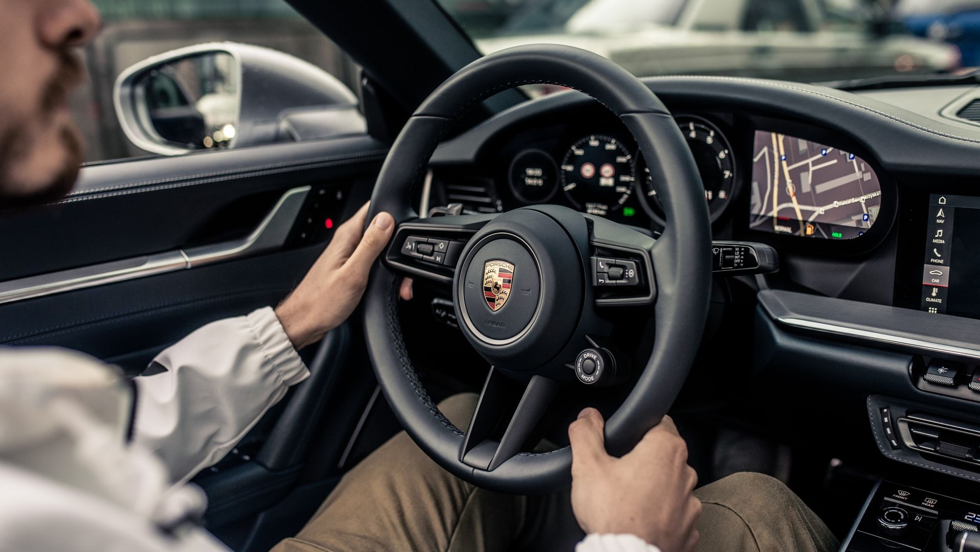 Wnętrze luksusowego samochodu Porsche, kierowca trzyma kierownicę, widoczny ekran nawigacji i deska rozdzielcza.