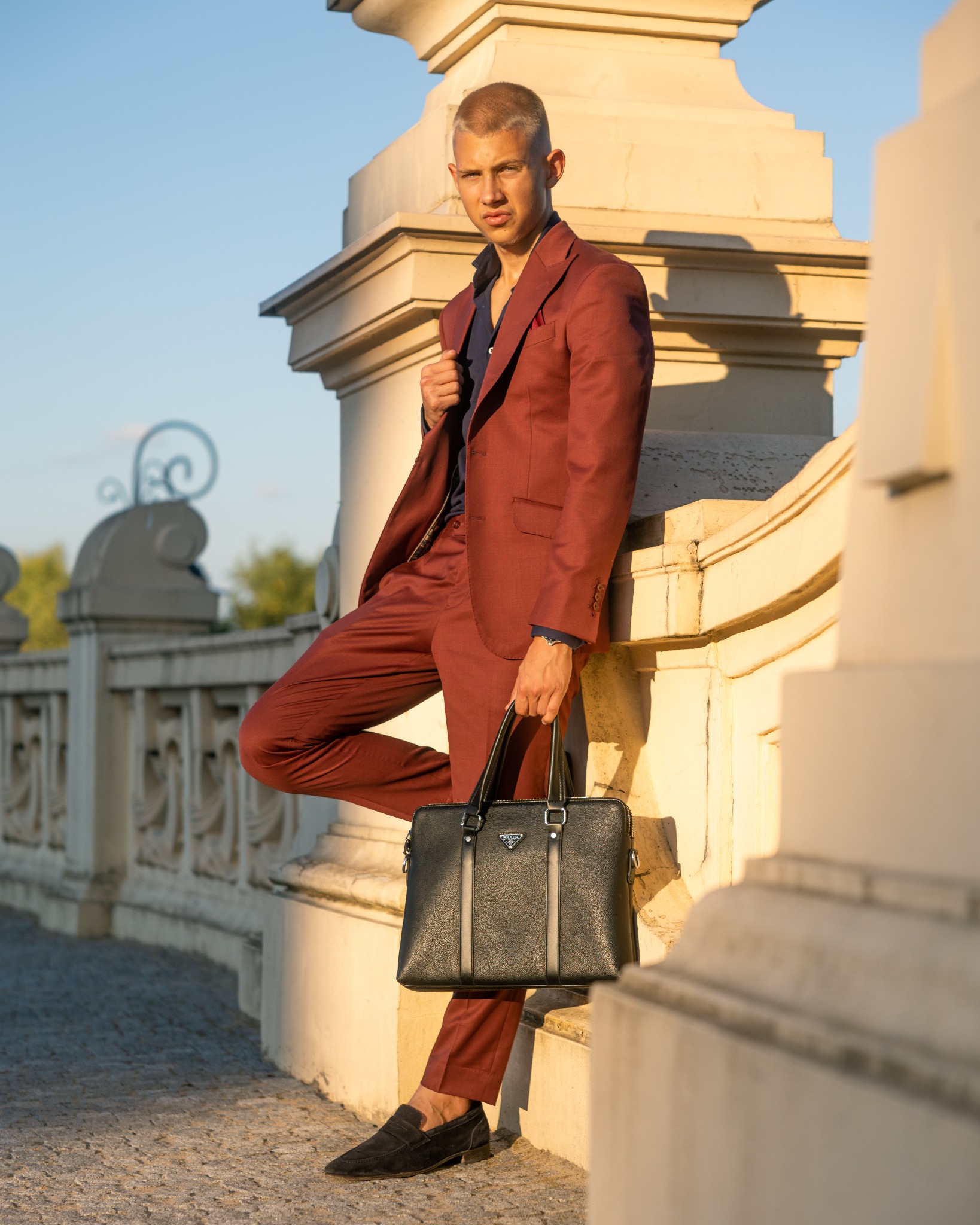 Mężczyzna w bordowym garniturze i czarnych mokasynach, oparty o kamienną balustradę, trzyma czarną skórzaną torbę Prada.