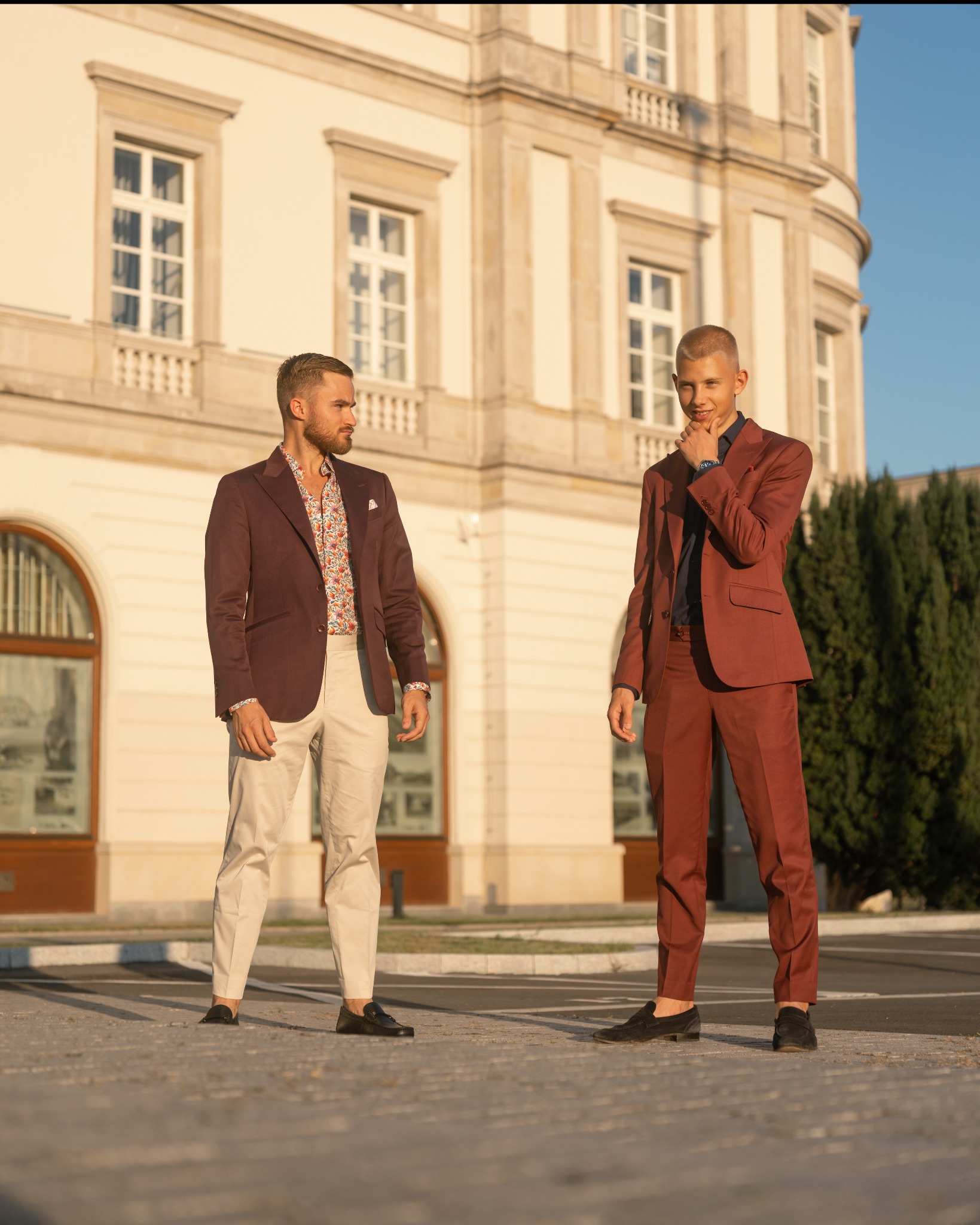 Dwóch mężczyzn w eleganckich, bordowych garniturach pozuje na tle klasycznej architektury, jeden z koszulą w kwiatowy wzór, drugi z ręką pod brodą.