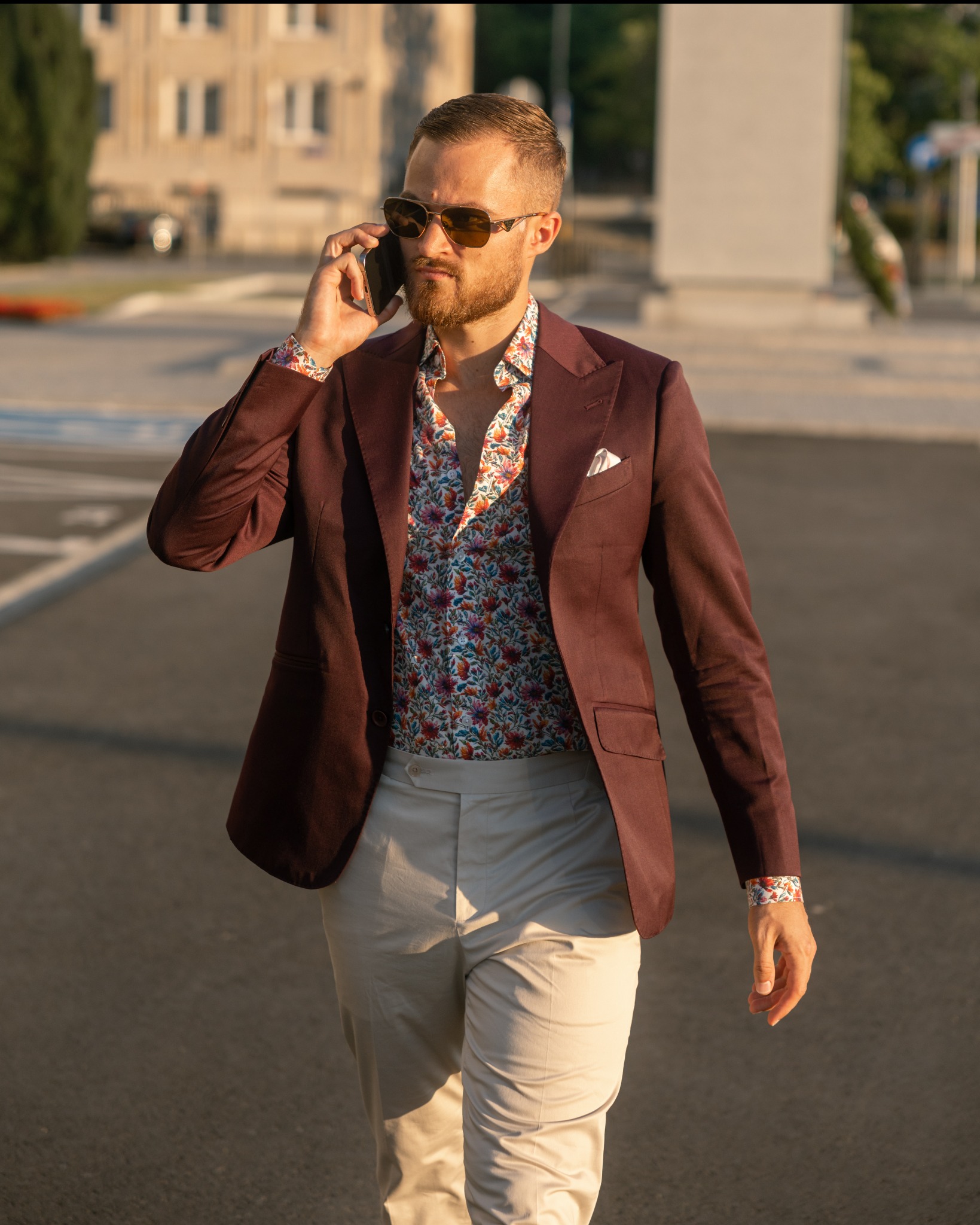 Mężczyzna w bordowym garniturze i kwiecistej koszuli rozmawia przez telefon na tle miejskiego krajobrazu w ciepłym świetle zachodzącego słońca.