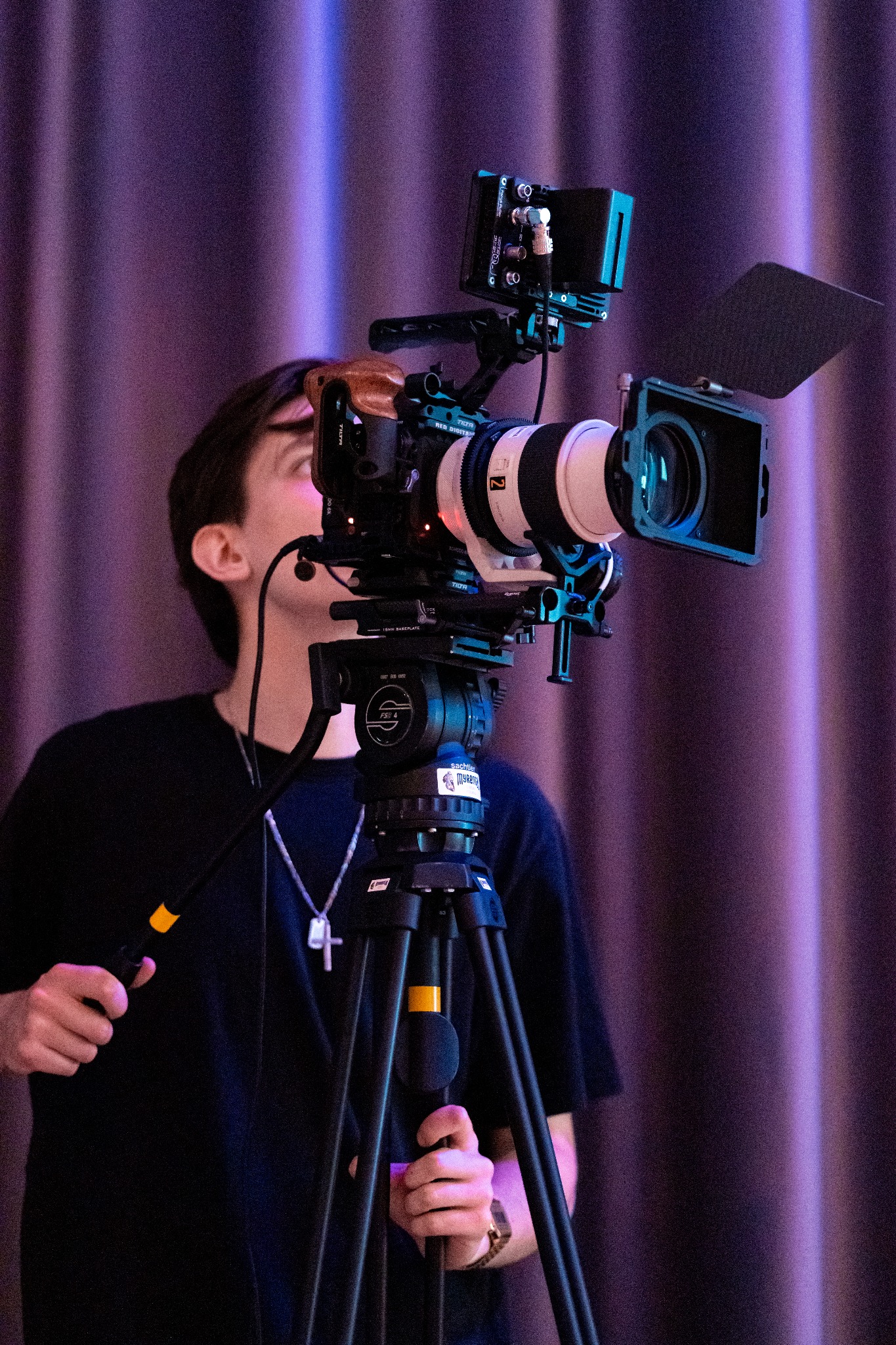 Kamerzysta obsługuje profesjonalną kamerę filmową na statywie, z monitorem i osłoną przeciwsłoneczną, ustawioną na tle fioletowej zasłony.