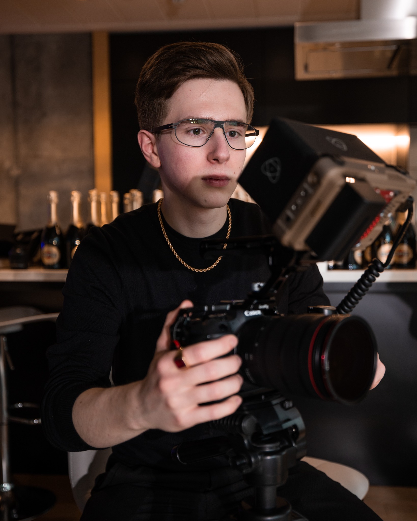 Młody mężczyzna w okularach i złotej biżuterii obsługuje profesjonalną kamerę Canon zamontowaną na statywie, z dodatkowym monitorem podglądowym; w tle butelki szampana.