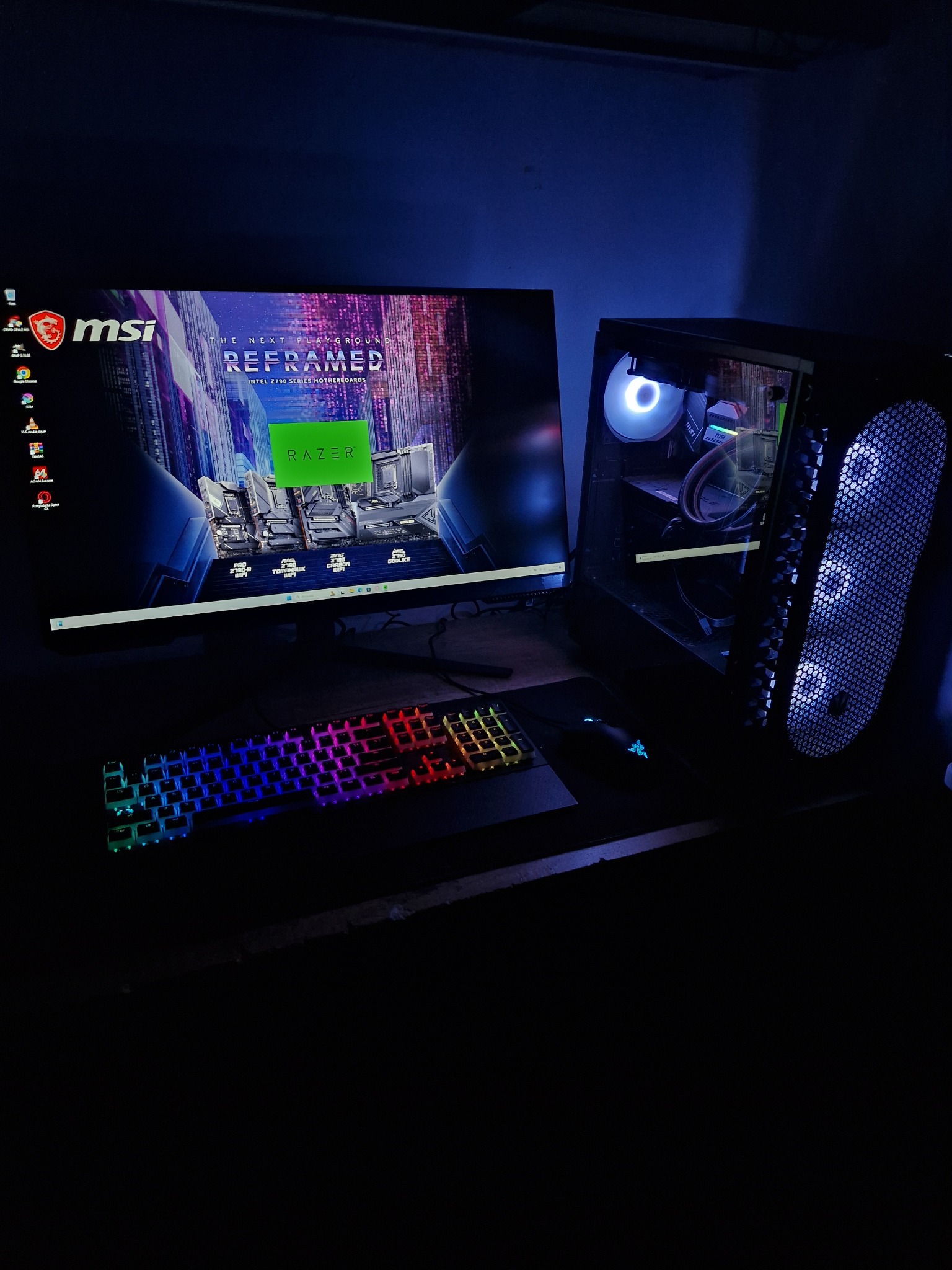 Komputer stacjonarny z podświetleniem RGB, monitor MSI wyświetlający reklamę płyty głównej, klawiatura i mysz Razer na biurku w ciemnym pokoju.