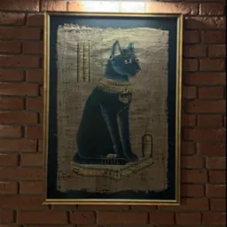 Oprawiony w złotą ramę obraz na papirusie przedstawia egipską boginię Bastet w formie czarnego kota z kolczykiem w nosie i bogatą biżuterią, wiszący na ścianie z czerwonej cegły pod lampą.