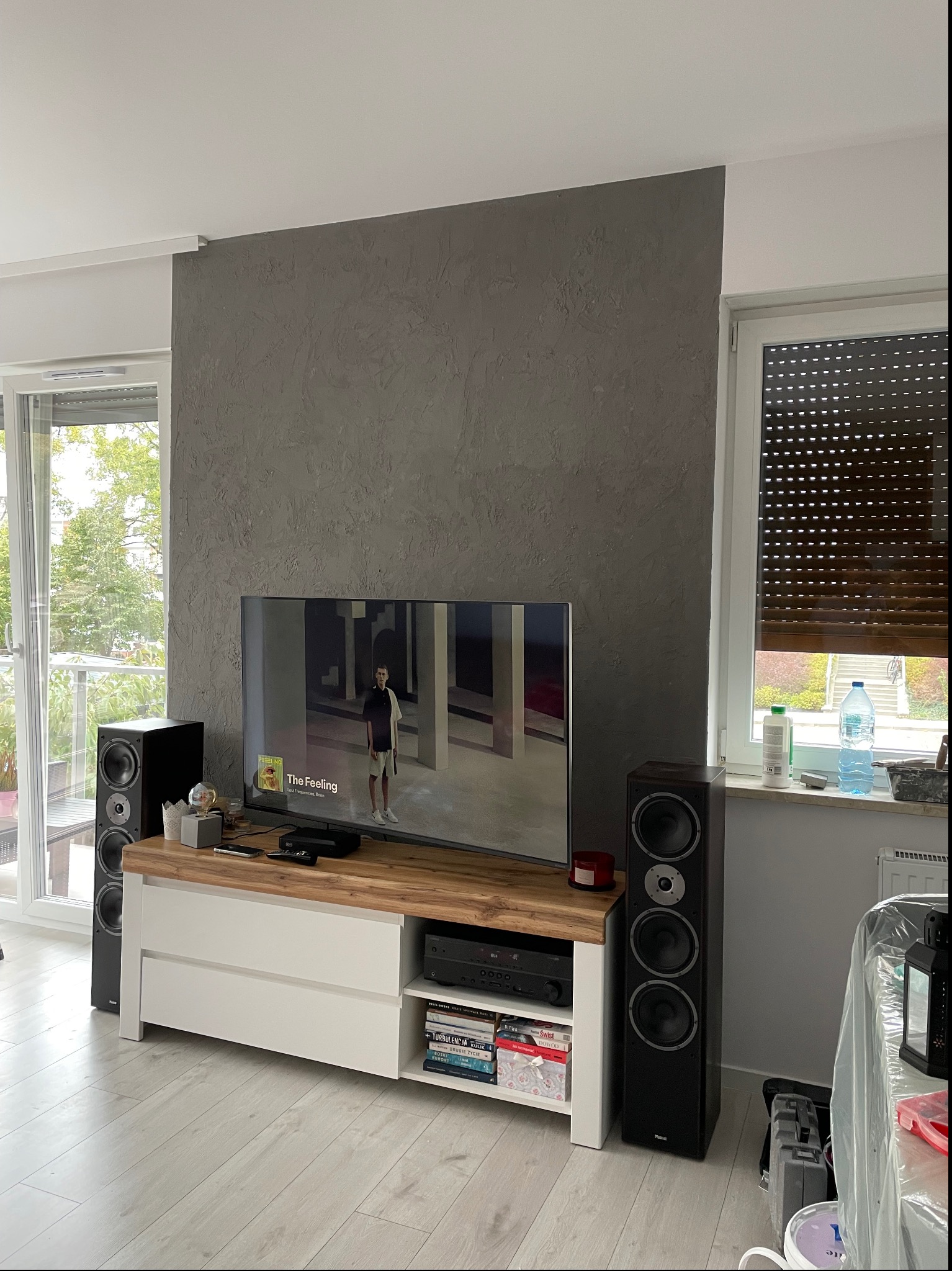 Szara ściana dekoracyjna o nierównej fakturze za telewizorem, obok okna z roletami, zestaw audio z kolumnami i szafką RTV.