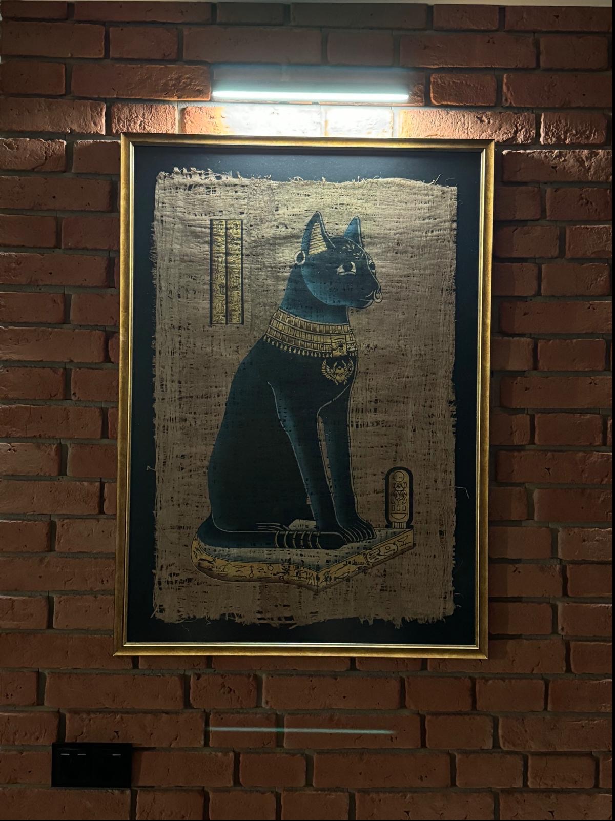 Oprawiony w złotą ramę obraz na papirusie przedstawia egipską boginię Bastet w formie czarnego kota z kolczykiem w nosie i bogatą biżuterią, wiszący na ścianie z czerwonej cegły pod lampą.