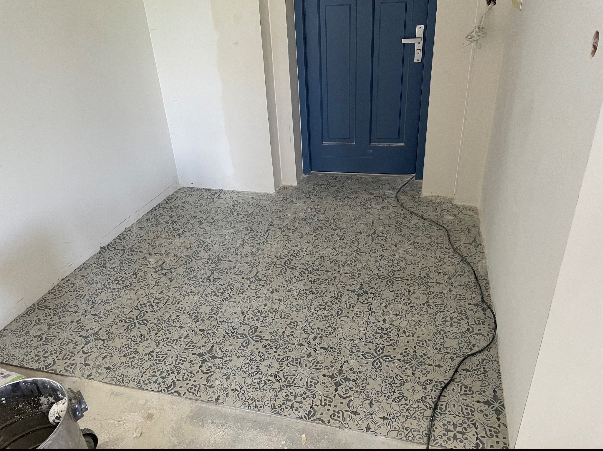 Ułożona podłoga z wzorzystych płytek w odcieniach szarości, widoczna niebieska stolarka drzwiowa i wiadro z zaprawą w rogu pomieszczenia, ściany przygotowane do malowania.