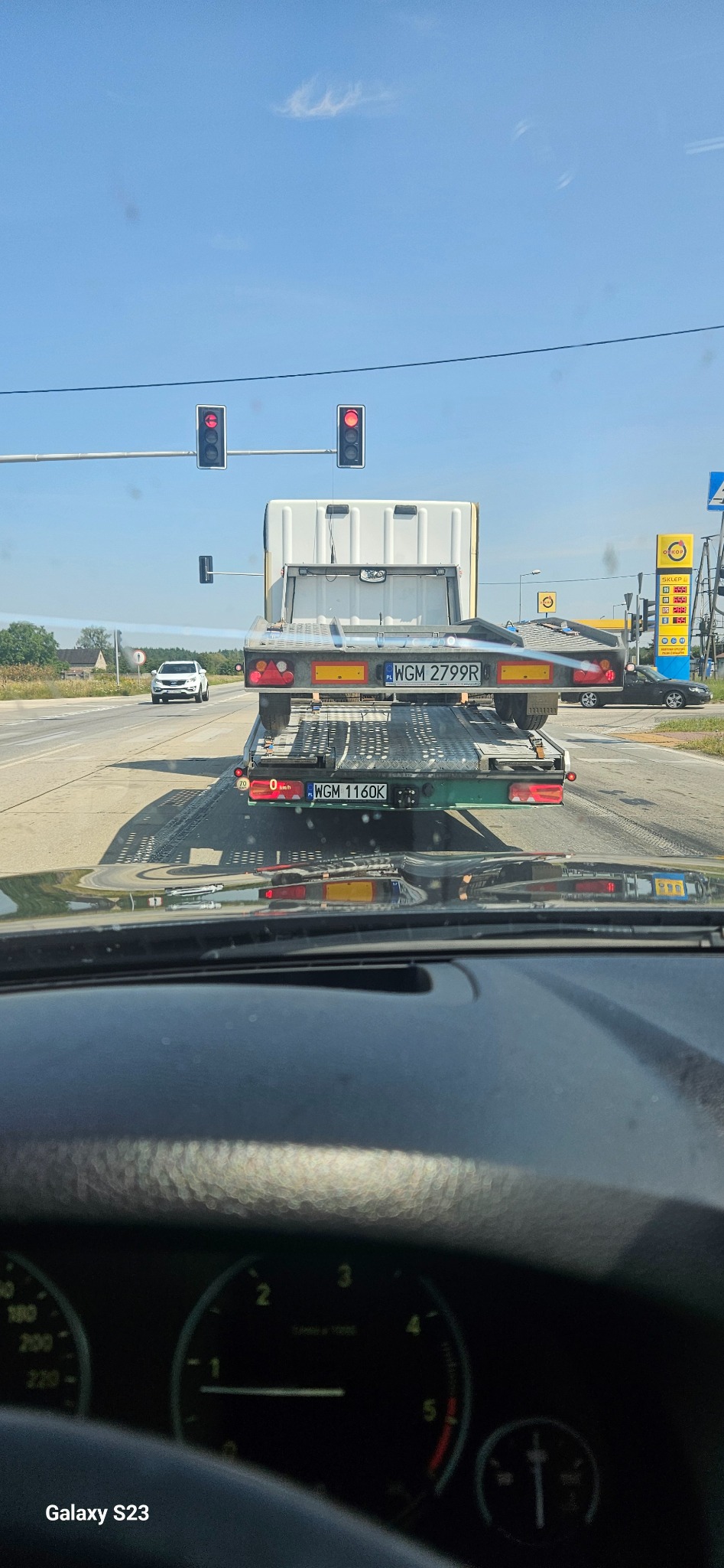 Widok z wnętrza samochodu na lawetę z przyczepą, stojącą na czerwonym świetle, z widocznym numerem rejestracyjnym WGM 2799R na przyczepie i WGM 1160K na lawecie, w tle stacja paliw Orlen...