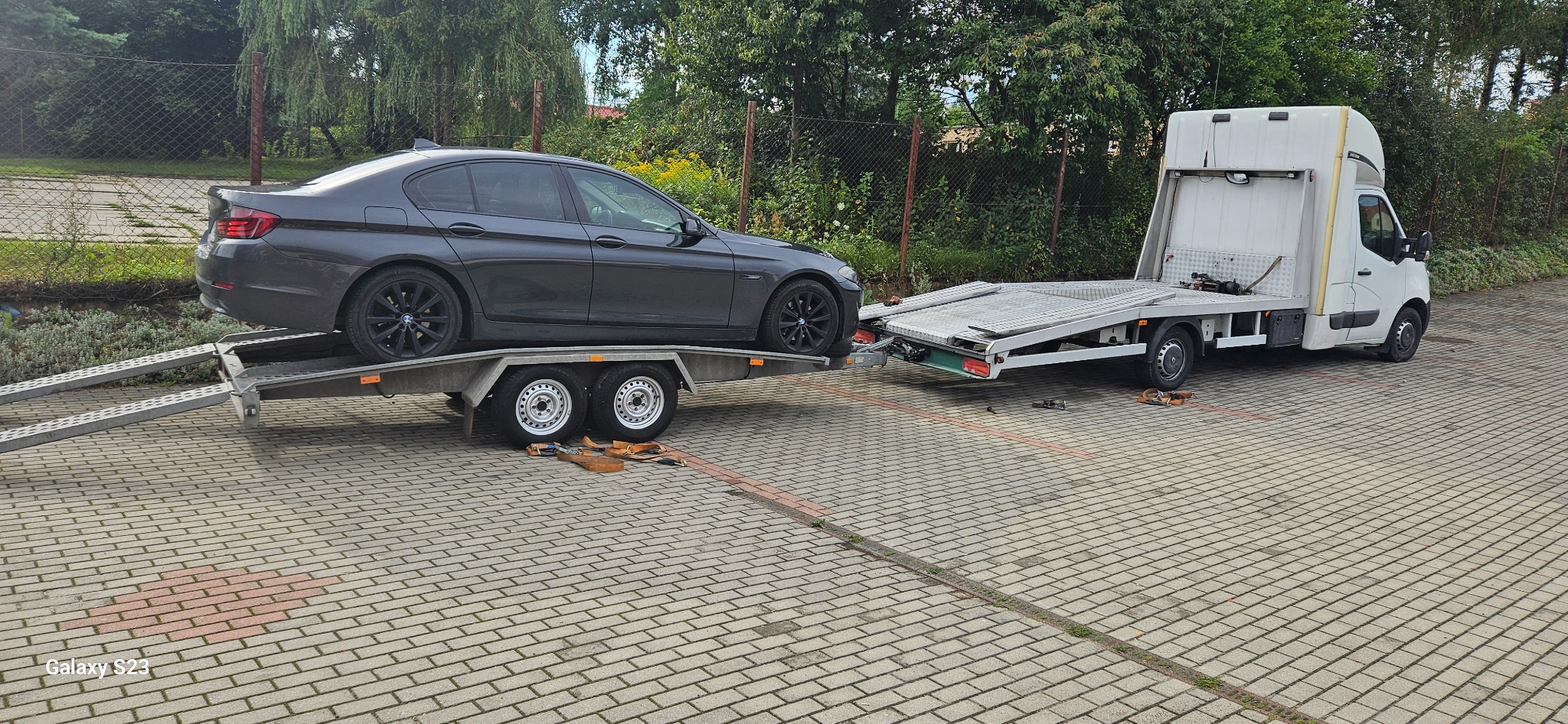 Szary samochód BMW na dwuosiowej przyczepie, podczepionej do białego pojazdu transportowego z zabudową, na brukowanym placu.