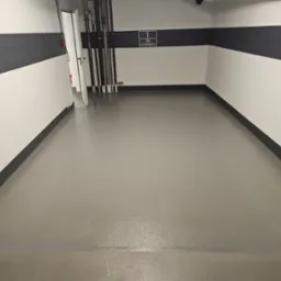 Szara, gładka podłoga w pomieszczeniu z biało-czarnymi ścianami. Widoczne rury przy ścianie i tabliczka z napisem 'TFG Asset Management'.