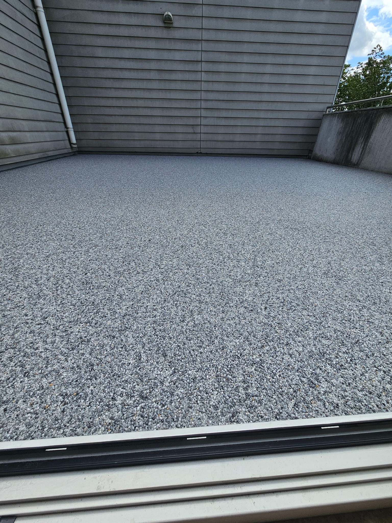 Szara, drobnoziarnista posadzka na balkonie, widok zza aluminiowej ramy okiennej. W tle szare deski elewacyjne i fragment nieba.