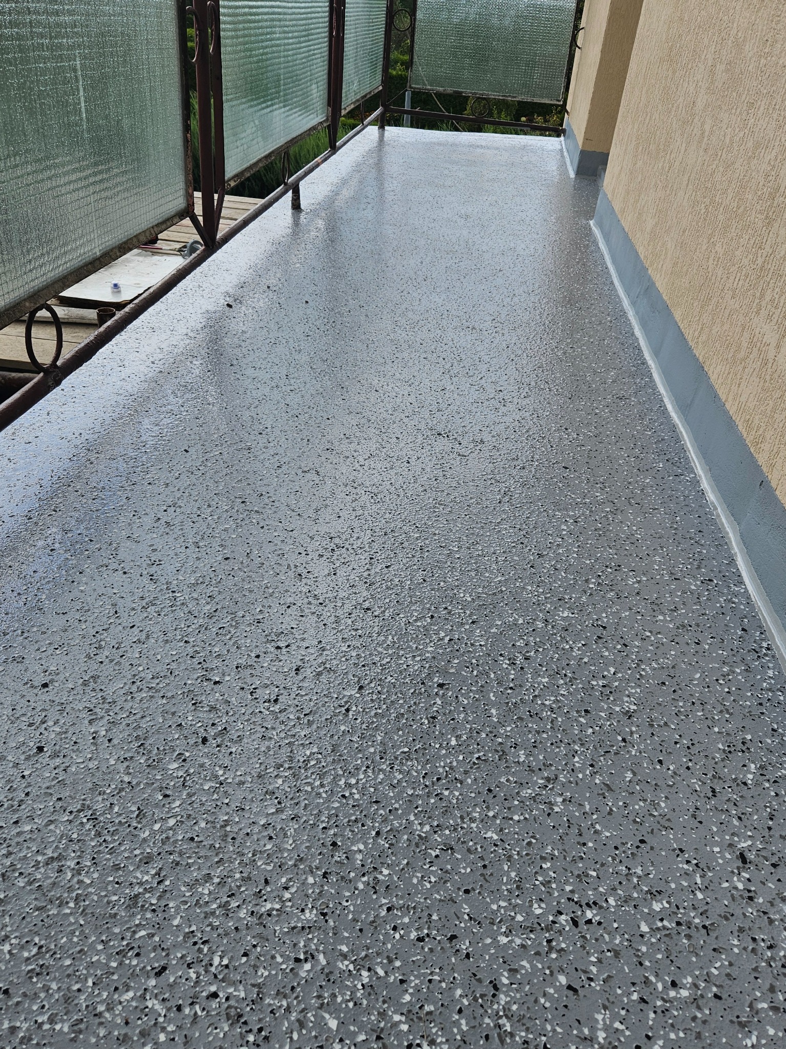 Szara, błyszcząca posadzka z drobnymi, czarnymi i białymi elementami na balkonie, z widoczną balustradą i fragmentem elewacji budynku.