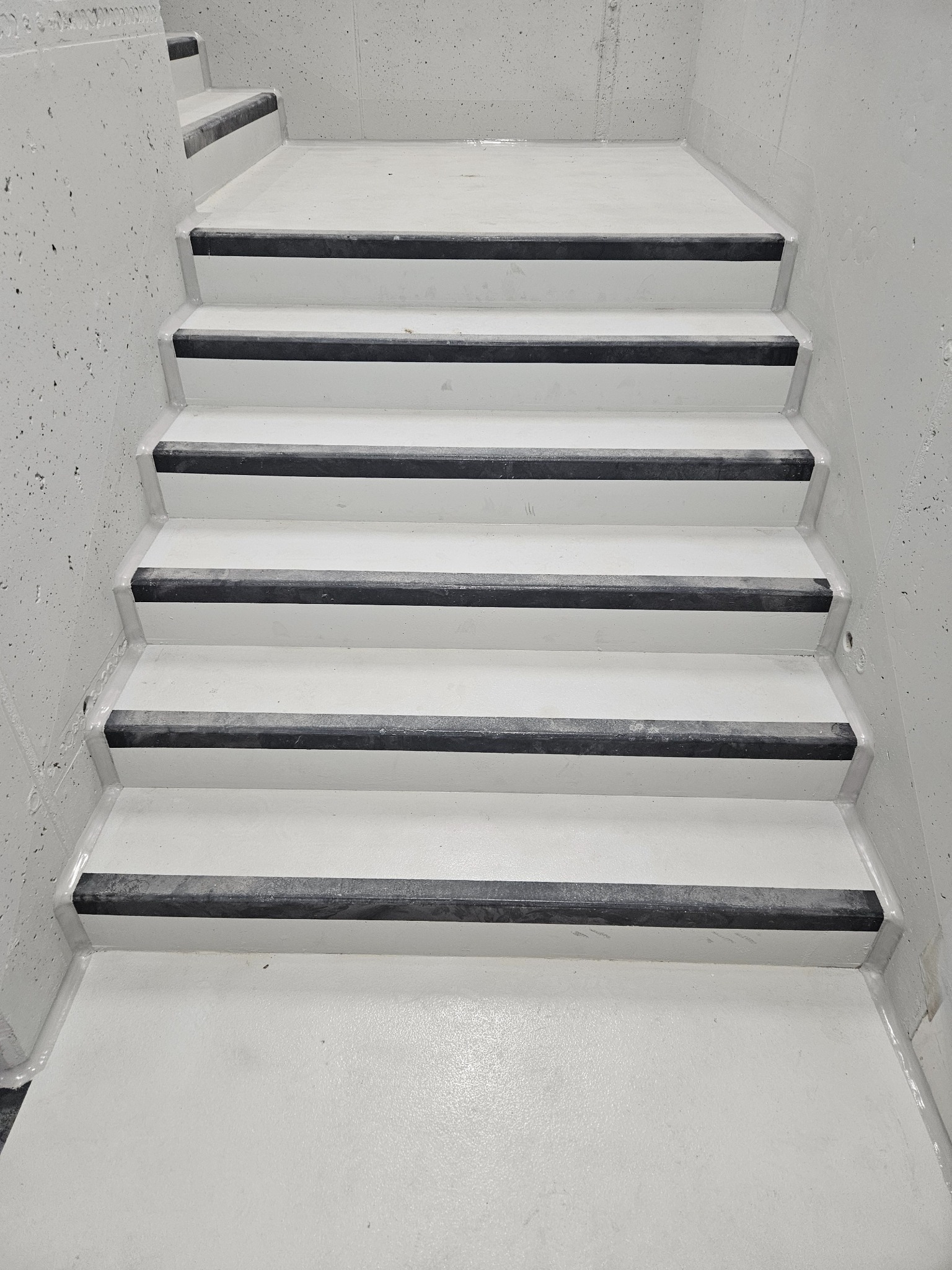 Białe schody w betonowej klatce schodowej z czarnymi, antypoślizgowymi paskami na krawędziach stopni, wykończone jednolitą, gładką powierzchnią.