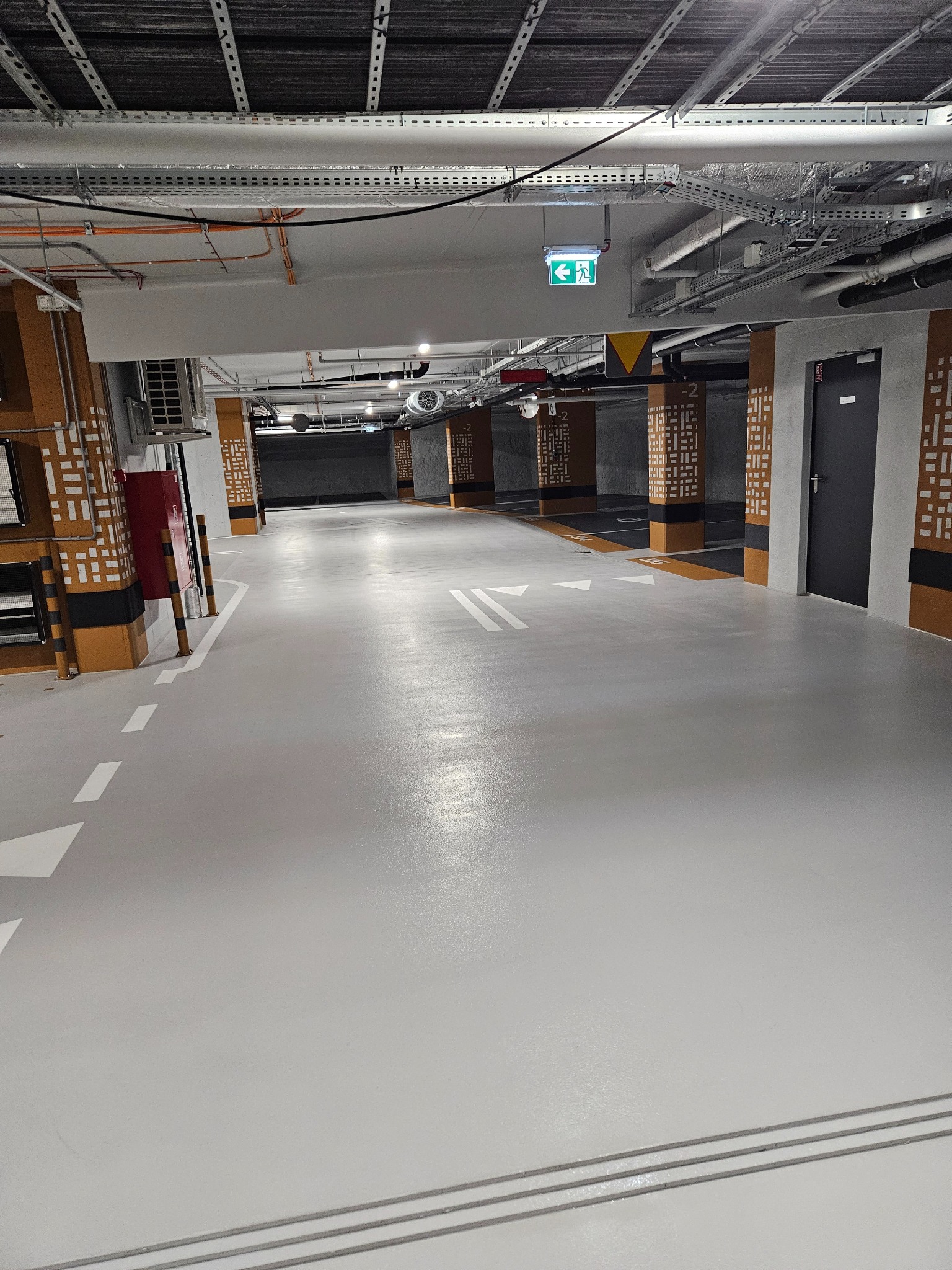 Jasnoszara, gładka nawierzchnia parkingu podziemnego z białymi znakami poziomymi. Widoczne kolumny z dekoracyjnym wzorem i instalacje pod sufitem.