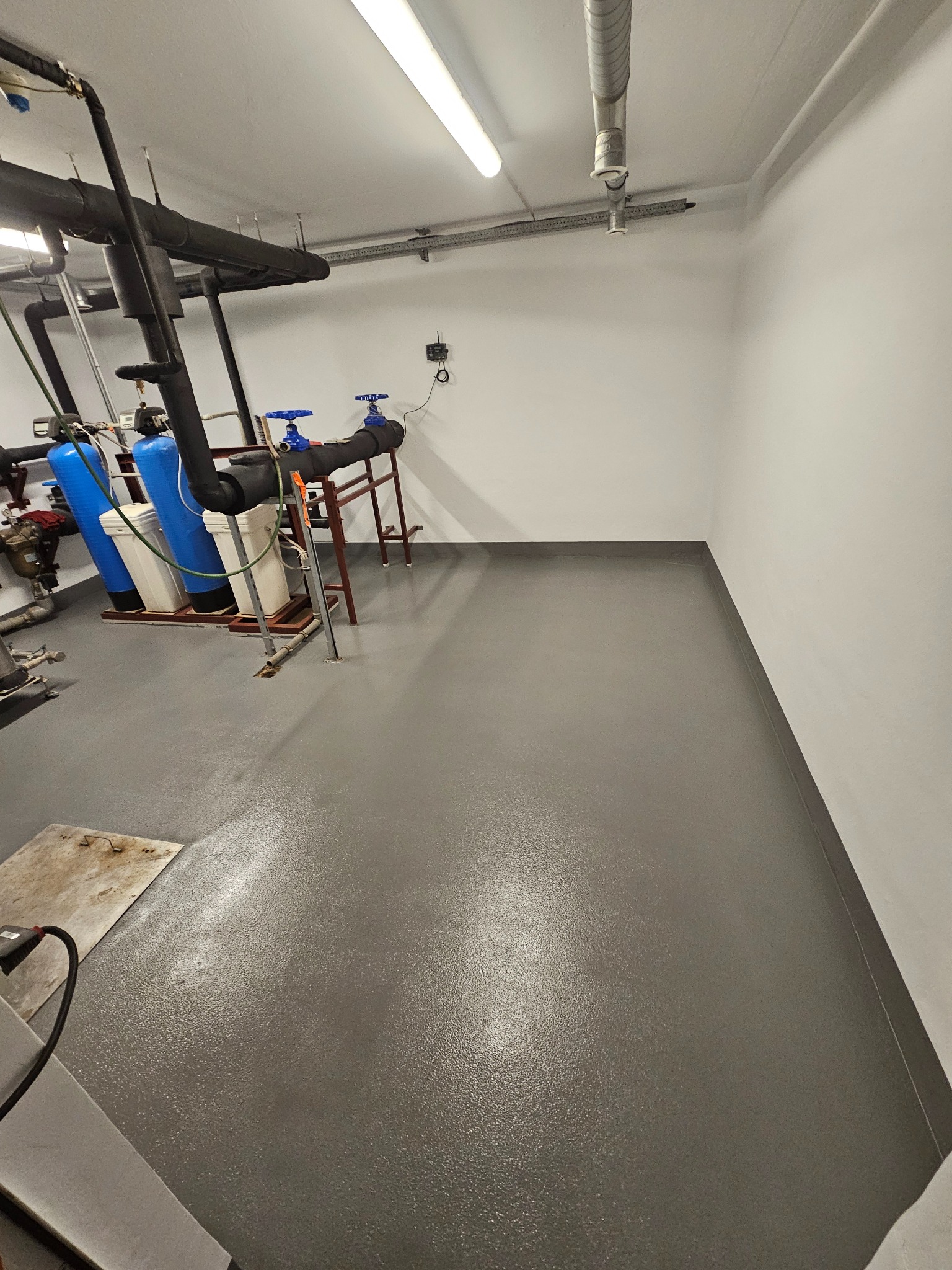 Szara, gładka powierzchnia w pomieszczeniu technicznym z instalacjami. Błyszcząca, jednolita posadzka w industrialnym wnętrzu. Widoczne rury i filtry wody.