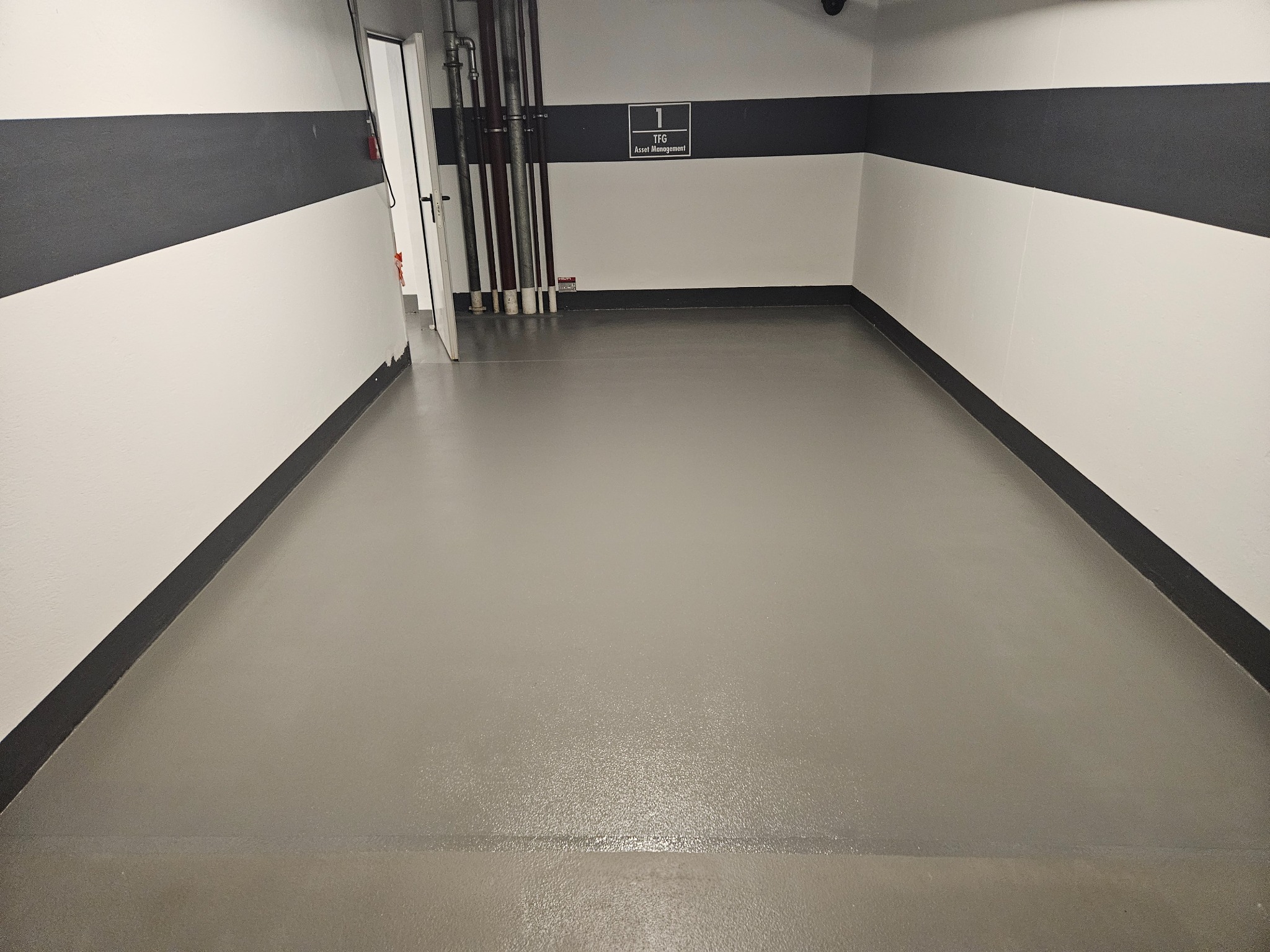 Szara, gładka podłoga w pomieszczeniu z biało-czarnymi ścianami. Widoczne rury przy ścianie i tabliczka z napisem 'TFG Asset Management'.