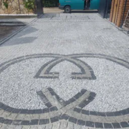 Ułożona nawierzchnia z kostki granitowej w odcieniach szarości z dekoracyjnym wzorem z jasnego żwiru i ciemniejszych kostek, widok z góry na wjazd do posesji.
