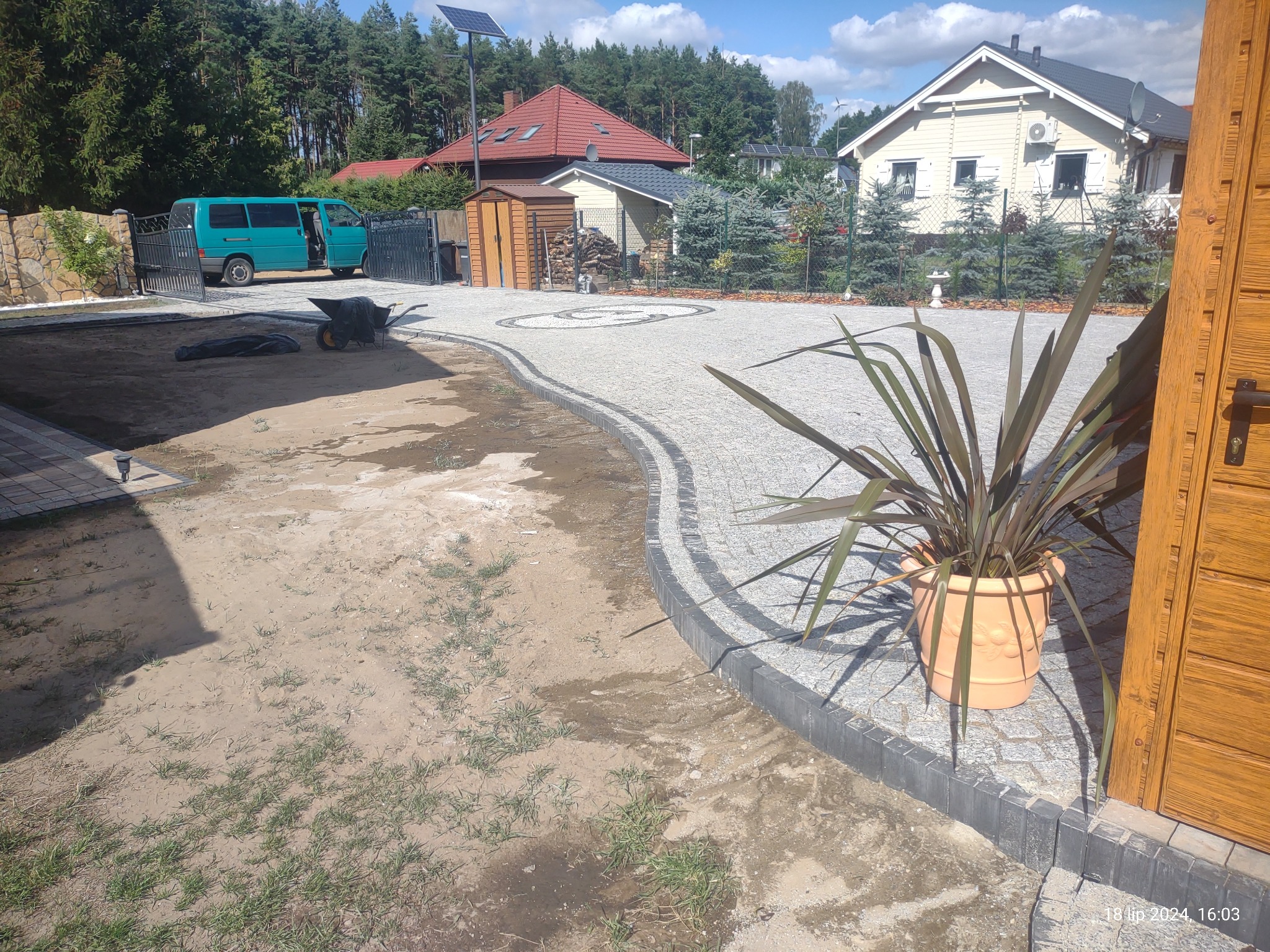 Układanie kostki granitowej w ogrodzie: widok na częściowo ułożoną nawierzchnię z kostki, z elementami dekoracyjnymi w kształcie okręgów, obok świeżo przygotowanego podłoża, z widocznym transportem...