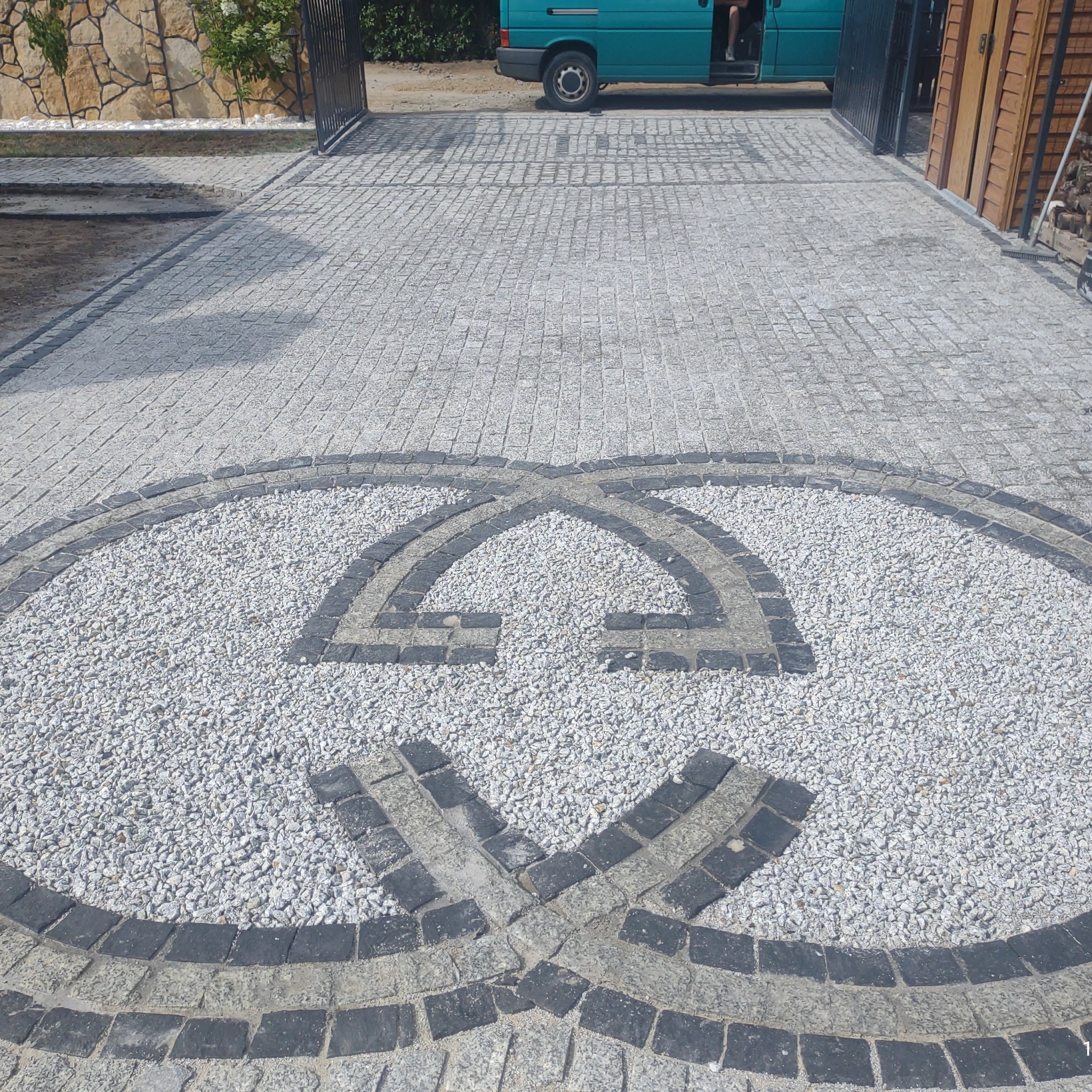 Ułożona nawierzchnia z kostki granitowej w odcieniach szarości z dekoracyjnym wzorem z jasnego żwiru i ciemniejszych kostek, widok z góry na wjazd do posesji.