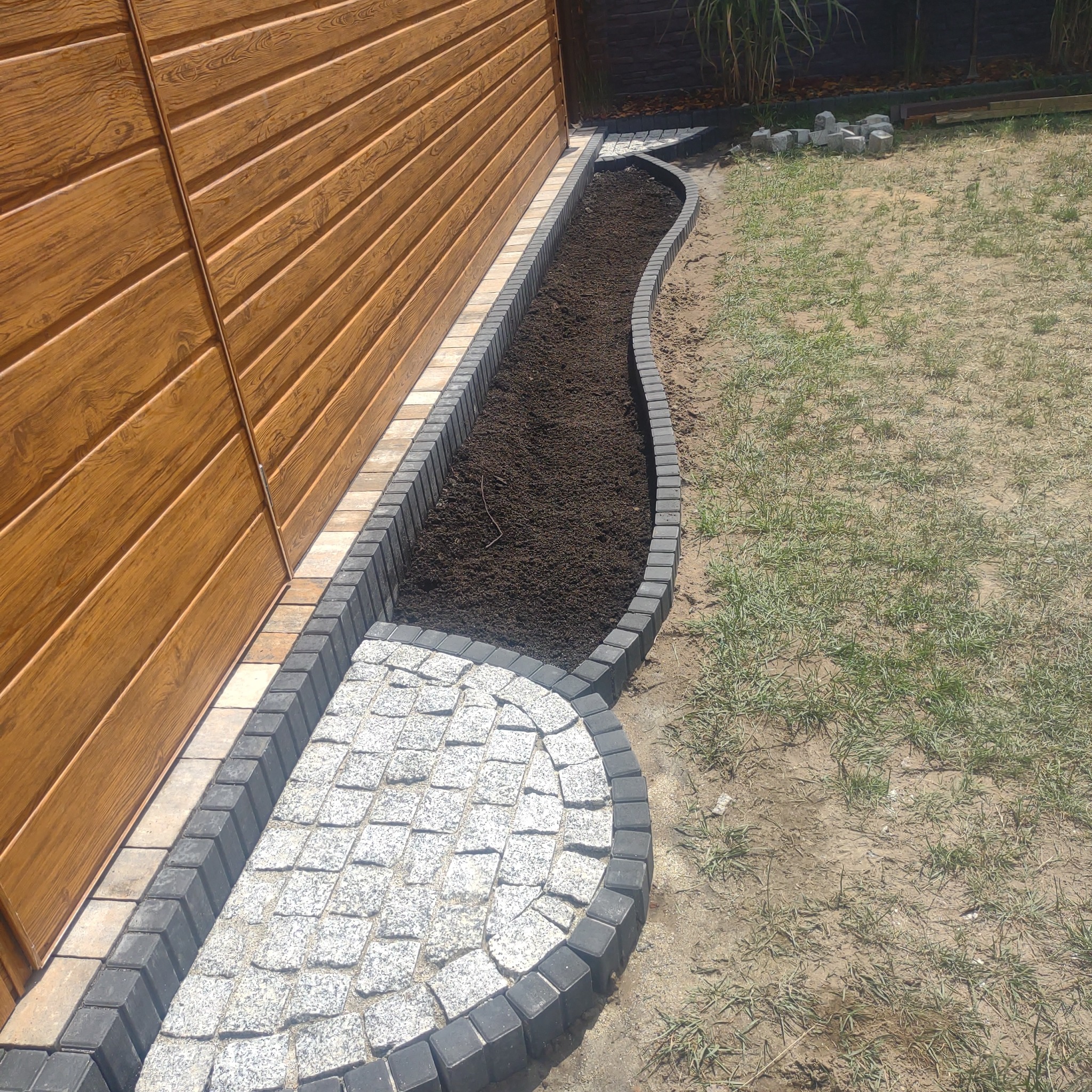 Obrzeże z kostki granitowej oddzielające rabatę z ziemią od trawnika, przy drewnianym ogrodzeniu.