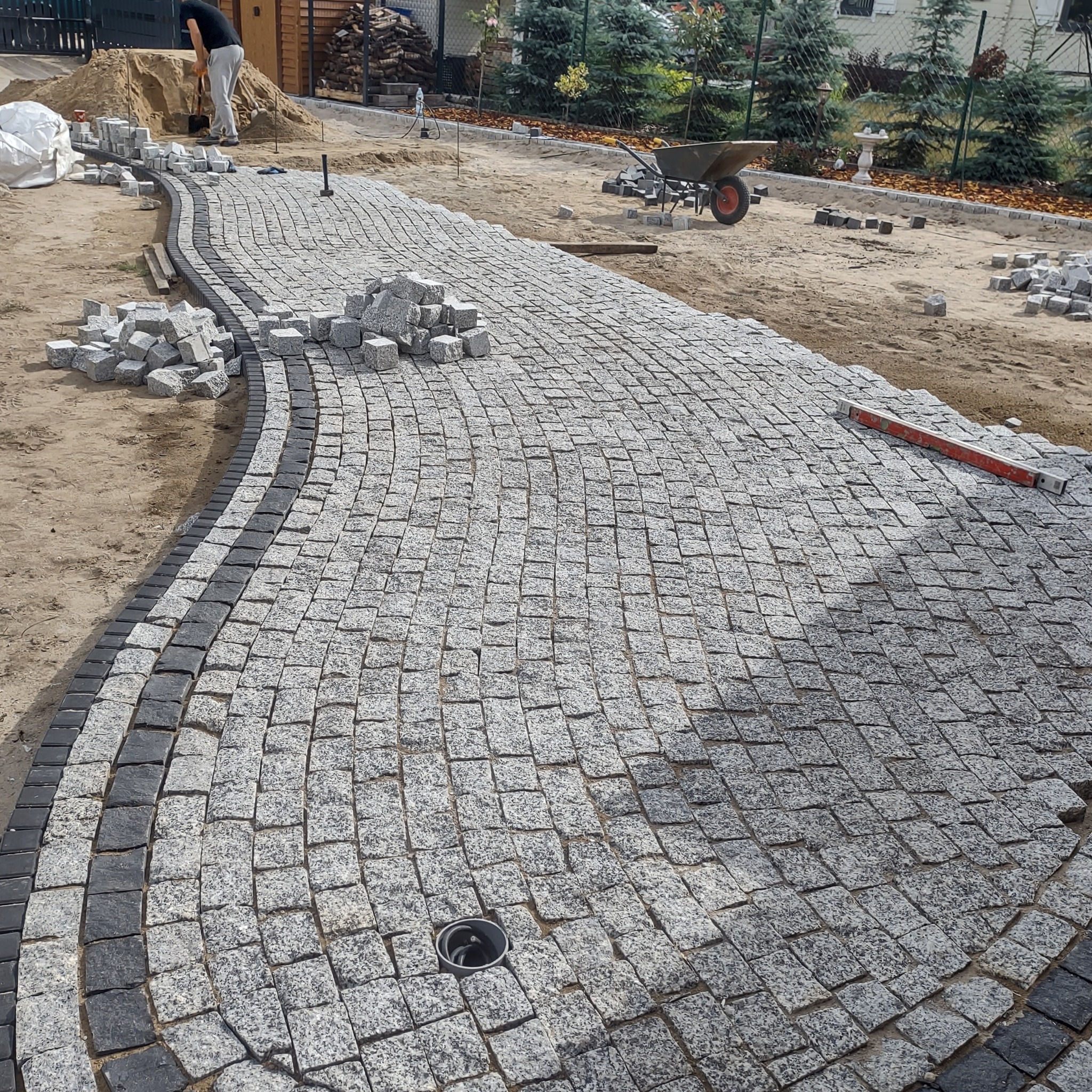 Fragment układanej ścieżki z kostki granitowej, z widocznym drenażem w postaci studzienki, w tle pracownik z łopatą i przygotowane materiały.