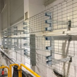 ELTREND Łukasz Balcerek - Metalowe korytka kablowe zamontowane na ścianie w surowym wnętrzu, widoczne elementy instalacji i fragment podnośnika z żółtą barierką.