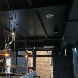 ELTREND Łukasz Balcerek - Czarne wnętrze lokalu z widocznymi lampami wiszącymi o różnym designie, zamontowanymi na szynoprzewodach pod sufitem. W tle okno z widokiem na budynek z cegły i zieleń. Widoczny także fragment...