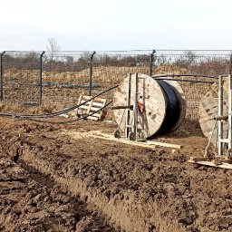 ELTREND Łukasz Balcerek - Prace ziemne związane z układaniem kabli elektrycznych: pracownik w kamizelce odblaskowej rozwija kabel z bębna, widoczne dwa bębny z kablami na drewnianych podporach, wykopany rów w ziemi, w tle...