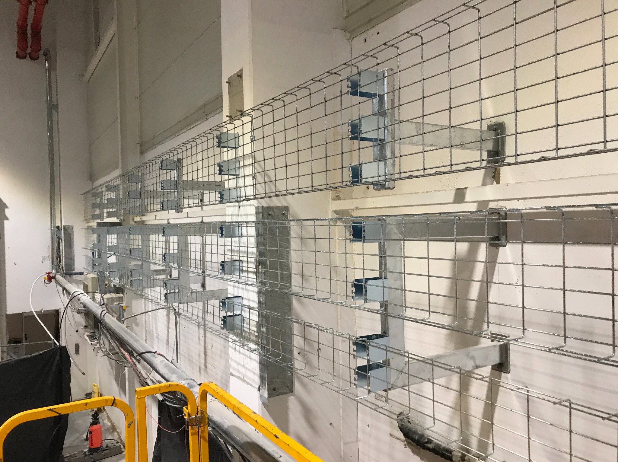 Metalowe korytka kablowe zamontowane na ścianie w surowym wnętrzu, widoczne elementy instalacji i fragment podnośnika z żółtą barierką.