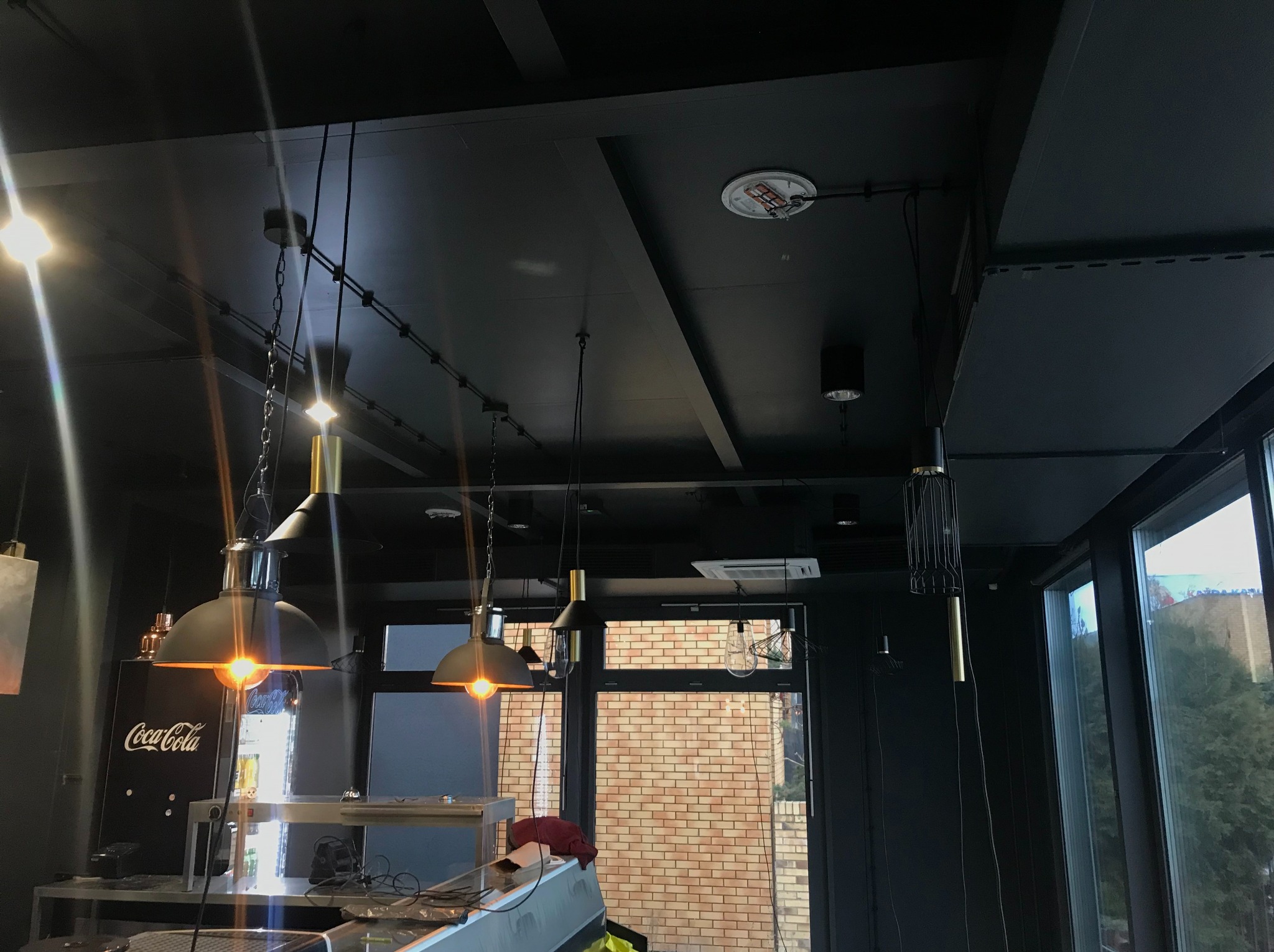 Czarne wnętrze lokalu z widocznymi lampami wiszącymi o różnym designie, zamontowanymi na szynoprzewodach pod sufitem. W tle okno z widokiem na budynek z cegły i zieleń. Widoczny także fragment...