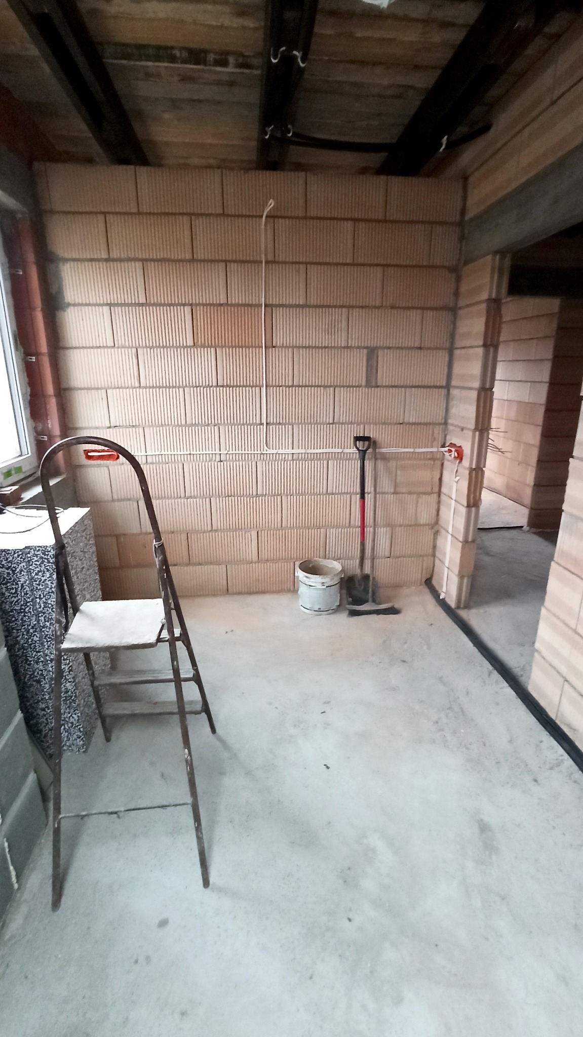 Surowy stan wnętrza w trakcie prac: widoczne przewody elektryczne wystające ze ściany z pustaków ceramicznych, drabina malarska, wiadro i łopata na podłodze.