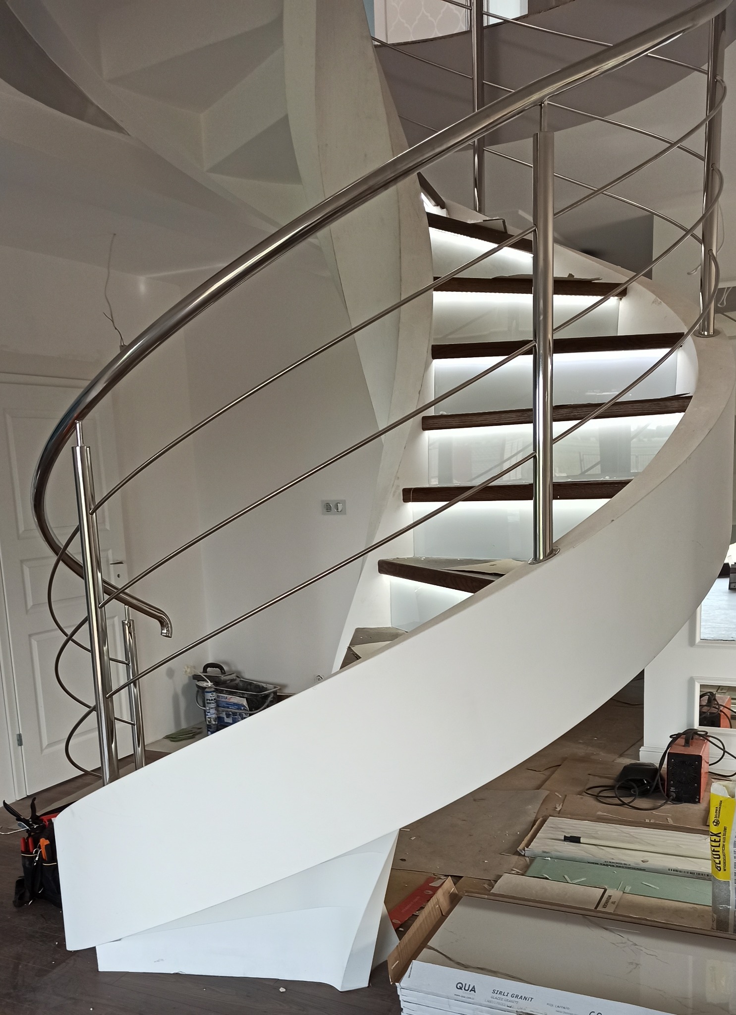Spiralne schody z oświetleniem LED na stopniach i chromowaną balustradą w trakcie montażu w Tomaszowie Mazowieckim, widoczne narzędzia i materiały budowlane.