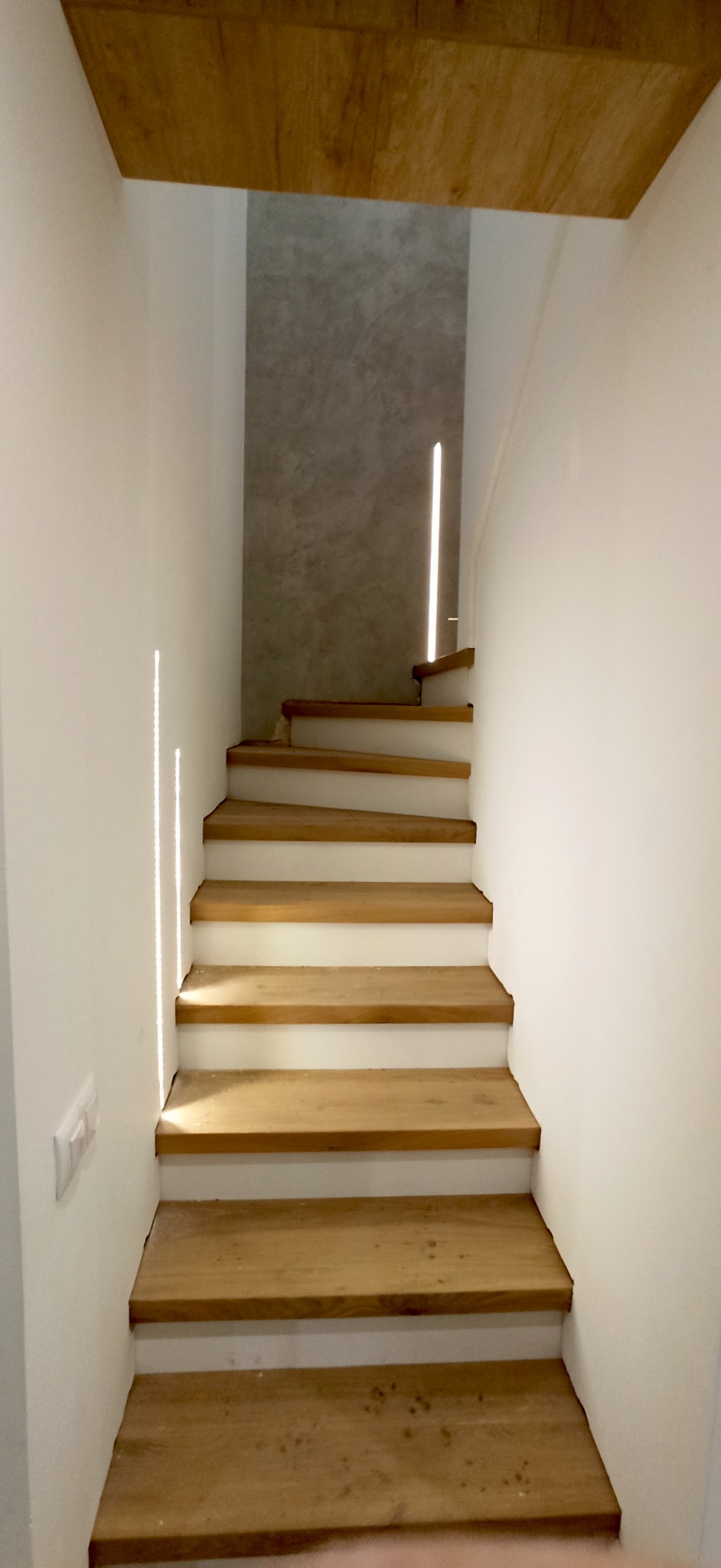 Nowoczesne schody drewniane z białymi podstopnicami, oświetlone naturalnym światłem wpadającym przez szczelinę w ścianie, widok z dołu na górę.