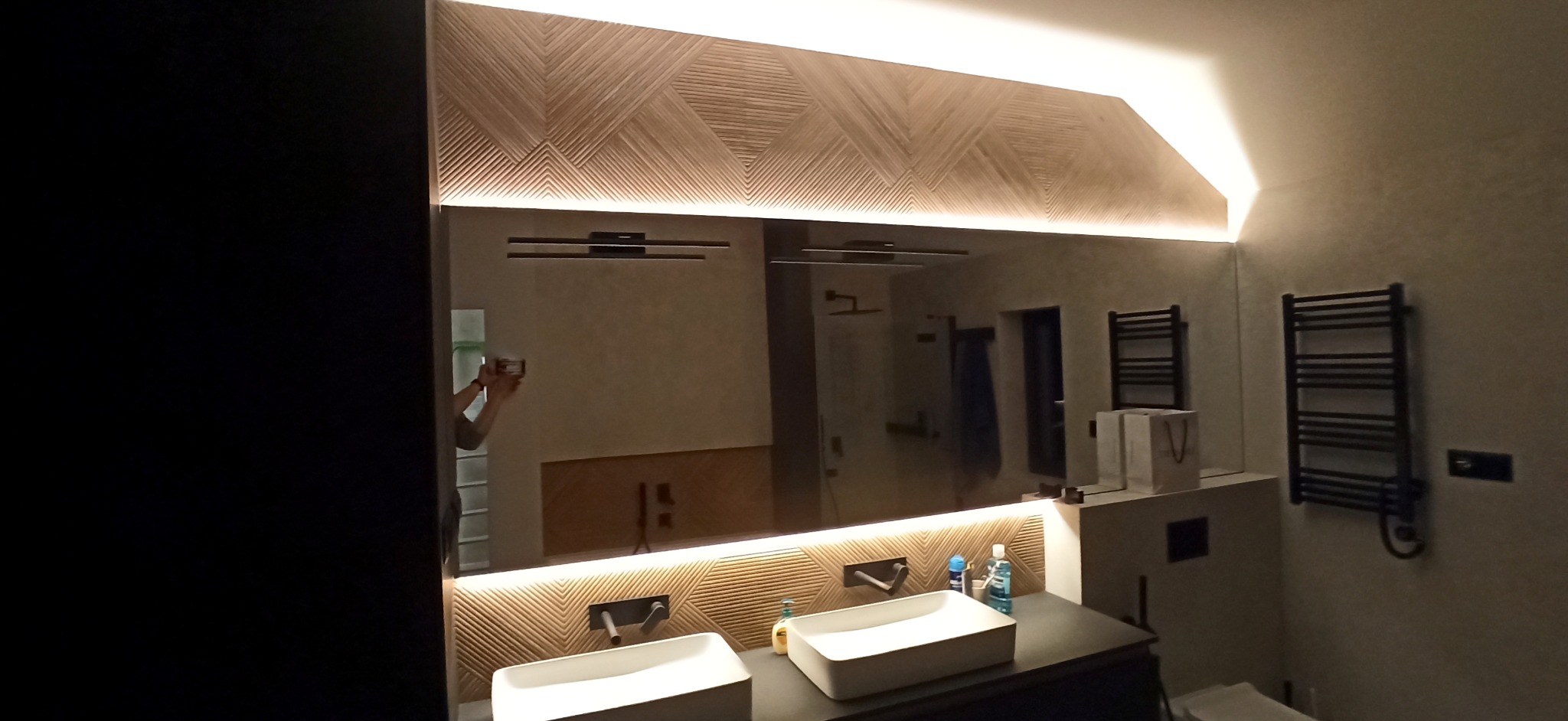 Nowoczesna łazienka z podświetlanym lustrem i dekoracyjnym panelem ściennym z drewna ułożonego we wzór chevron, widoczne dwie białe umywalki nablatowe i czarny grzejnik drabinkowy.