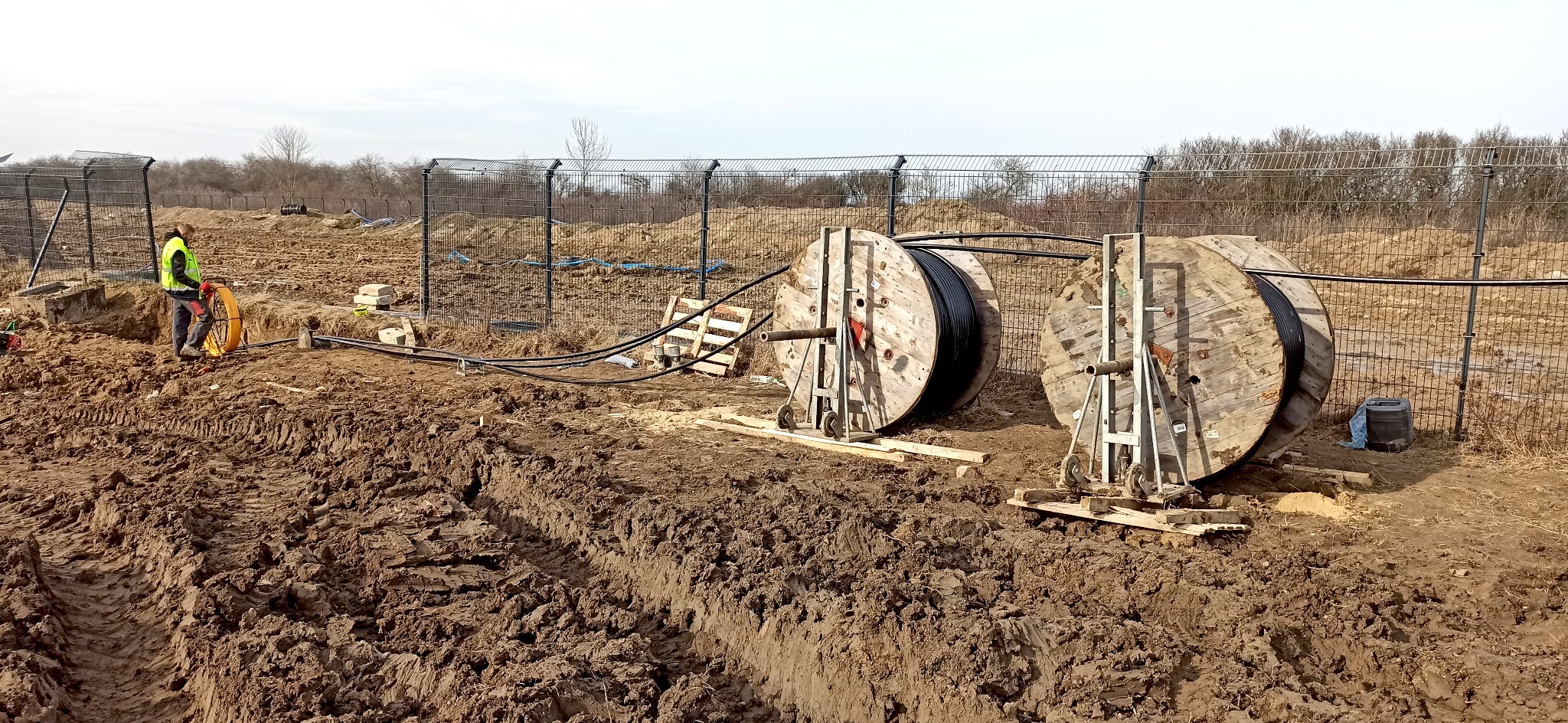 Prace ziemne związane z układaniem kabli elektrycznych: pracownik w kamizelce odblaskowej rozwija kabel z bębna, widoczne dwa bębny z kablami na drewnianych podporach, wykopany rów w ziemi, w tle...