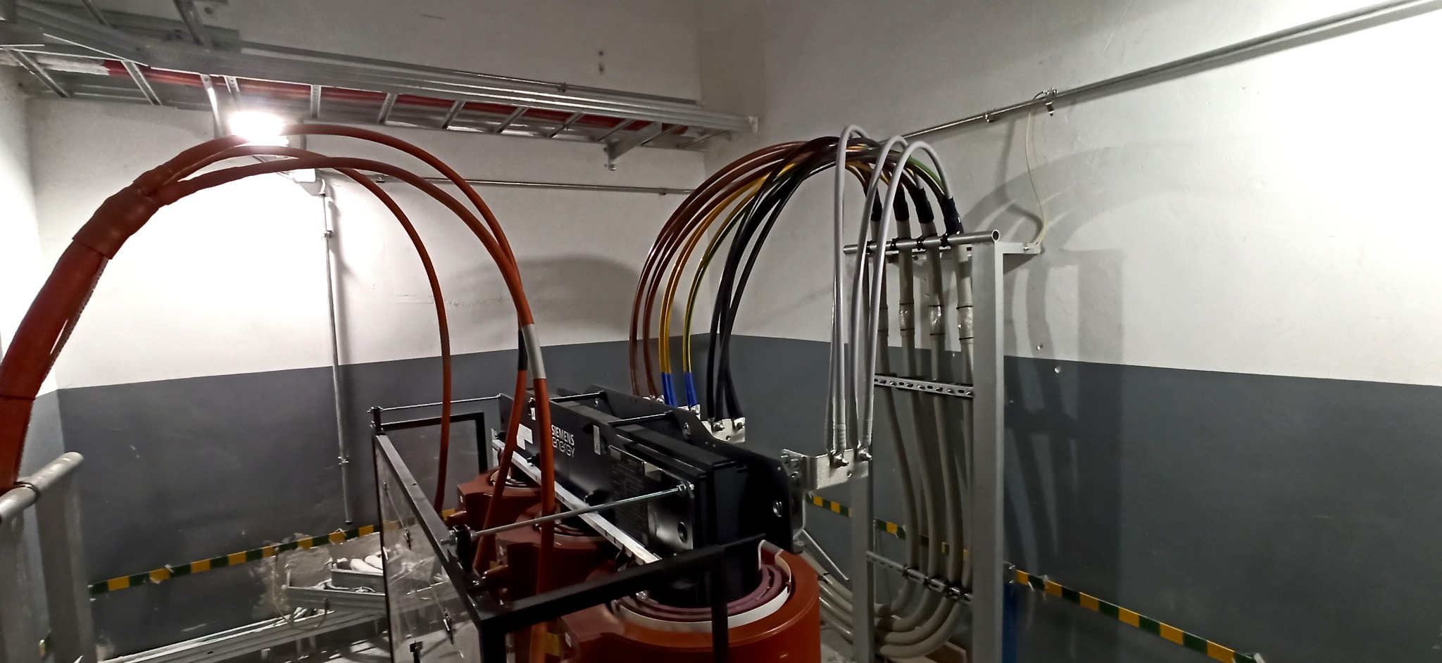 Wnętrze pomieszczenia z transformatorem Siemens Energy, widoczne kable o różnych kolorach prowadzące do urządzenia, oraz metalowe konstrukcje wsporcze na ścianach.