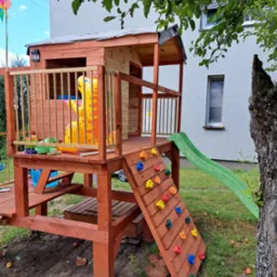 Drewniany plac zabaw z domkiem, zjeżdżalnią i ścianką wspinaczkową na tle trawnika i budynku mieszkalnego.