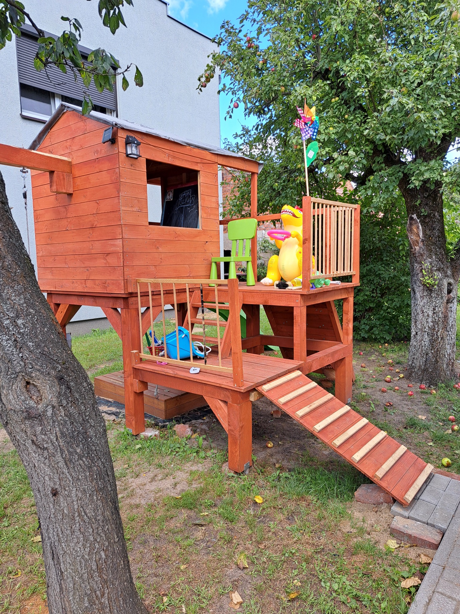 Drewniany plac zabaw z domkiem na palach, zjeżdżalnią i balustradą, zabawki na platformie, w tle drzewo owocowe z opadłymi jabłkami.