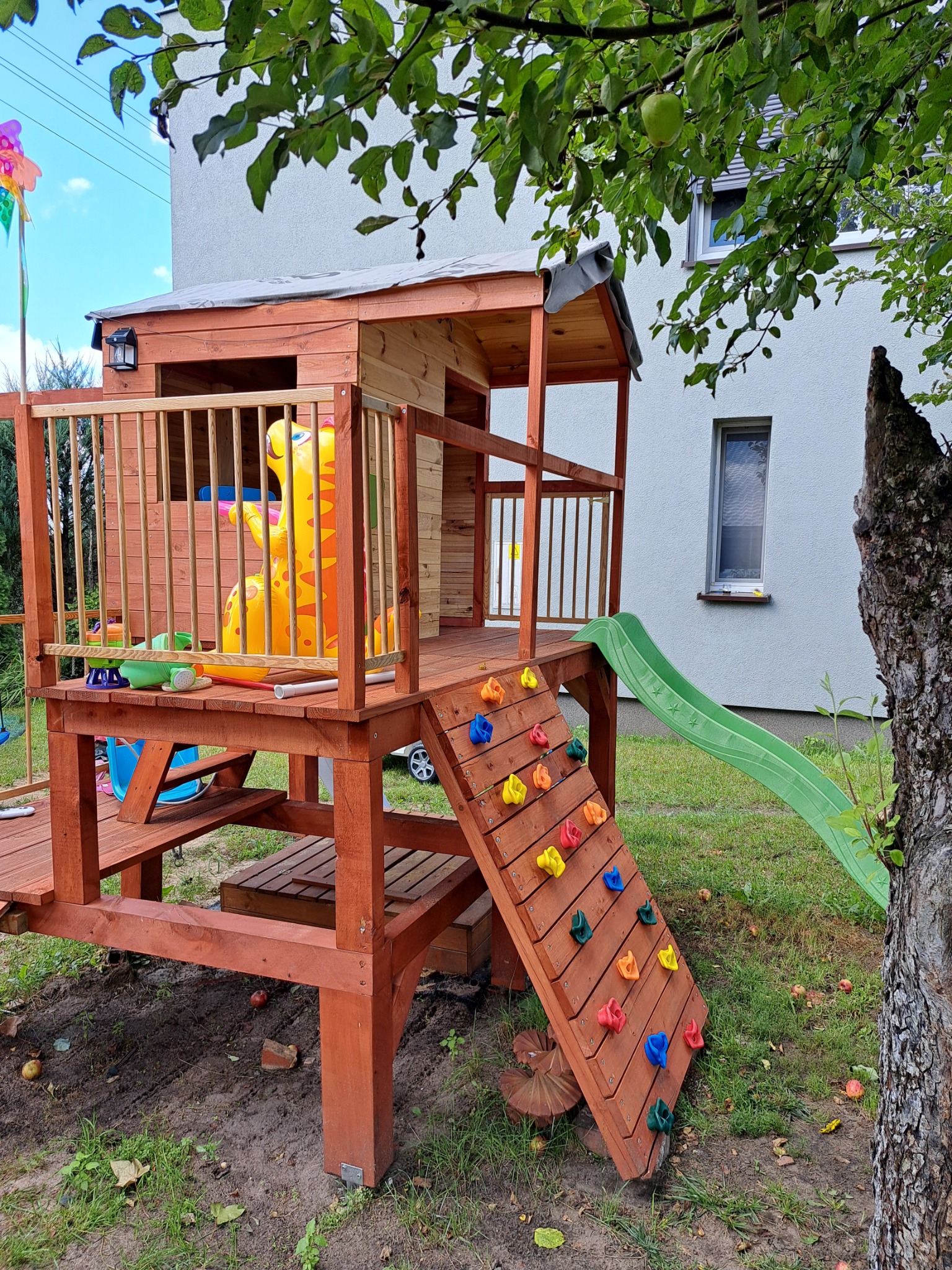 Drewniany plac zabaw z domkiem, zjeżdżalnią i ścianką wspinaczkową na tle trawnika i budynku mieszkalnego.