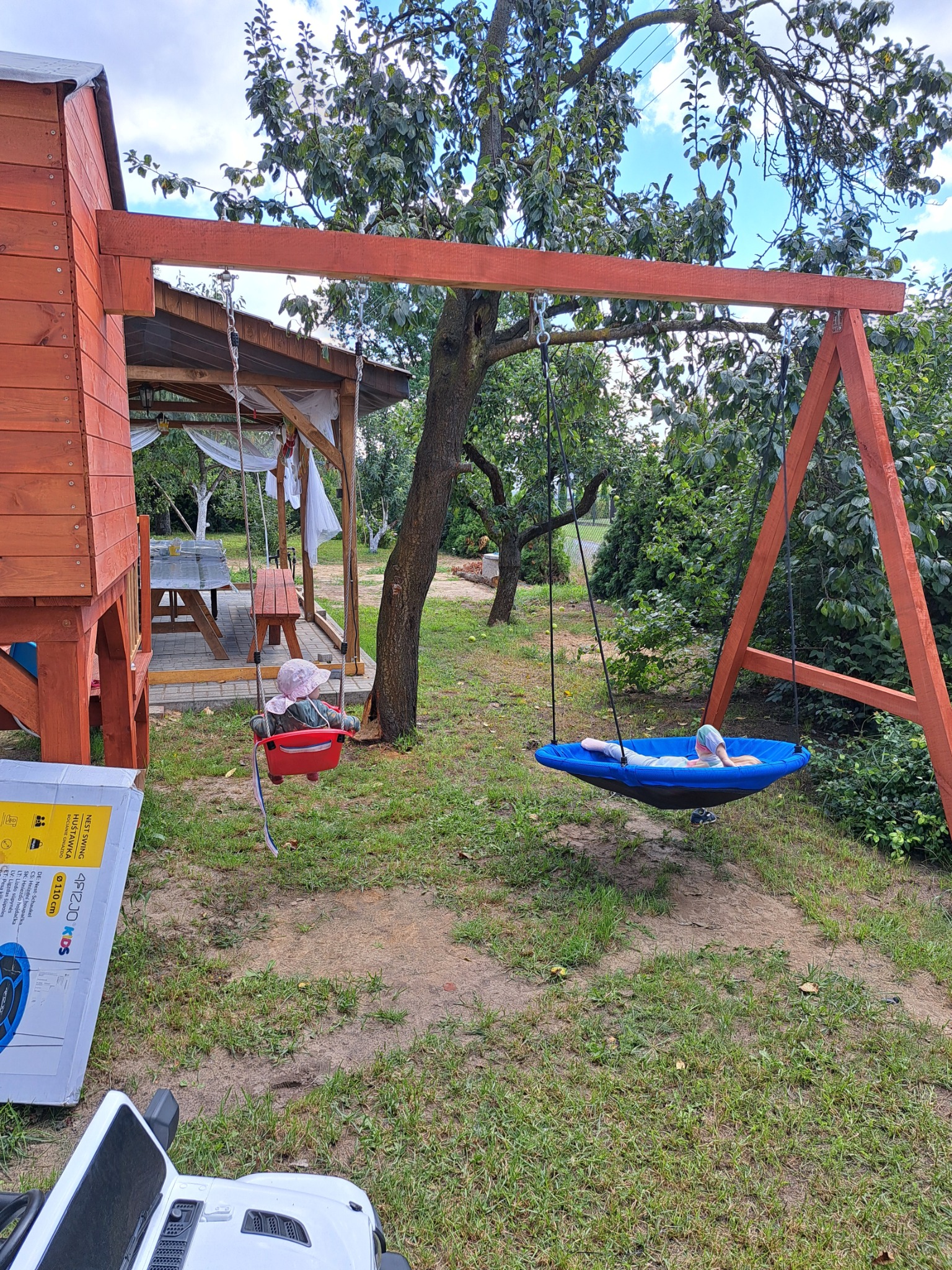 Dzieci na huśtawkach: jedno w czerwonym kubełku, drugie w niebieskim gnieździe, na tle drewnianej konstrukcji, domku i ogrodu.