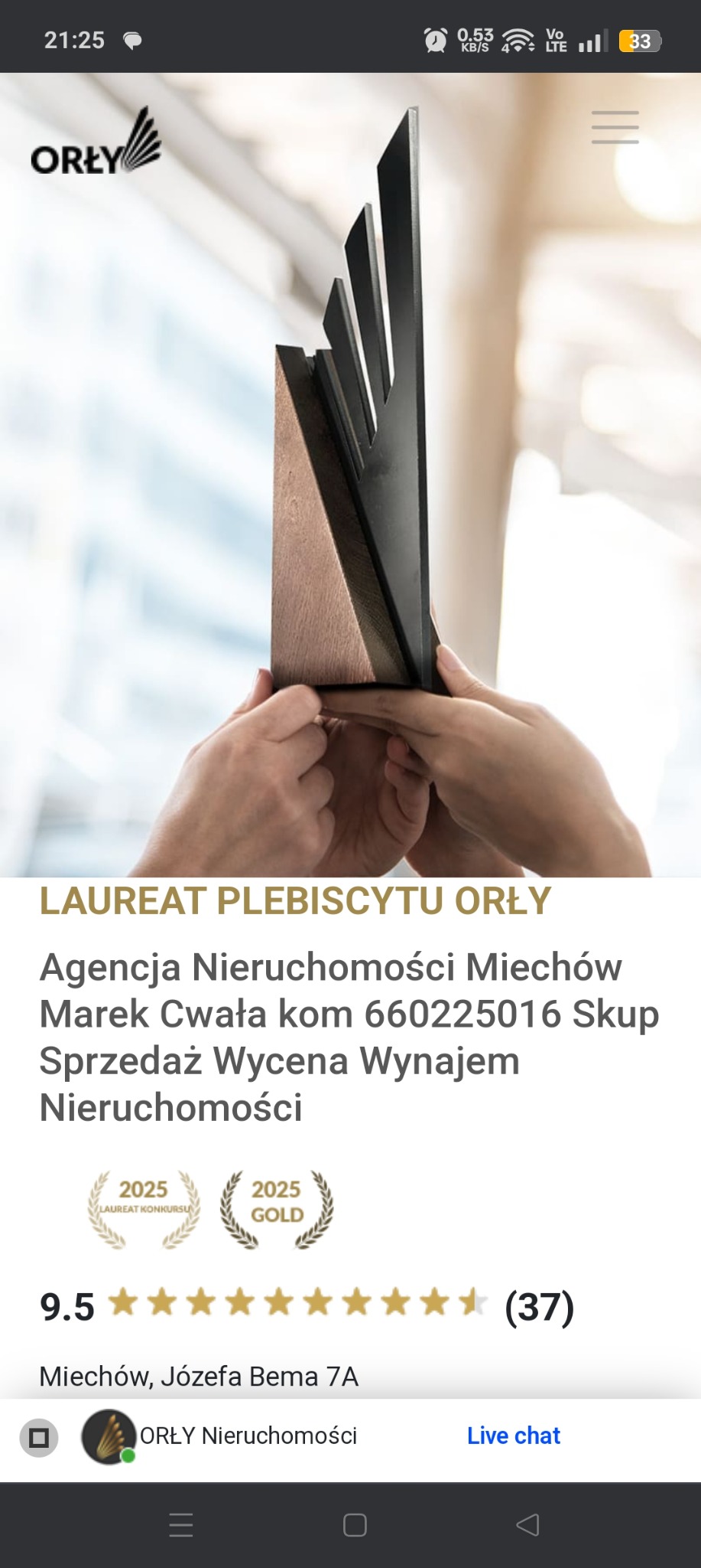 Dłonie trzymają drewniano-metalową statuetkę nagrody Orły, na rozmytym tle okna, w tle widoczny tekst reklamowy agencji nieruchomości z Miechowa.