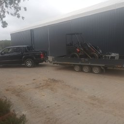 LASKOWSCY Roboty Ziemne Usługi Rolnicze Marcin Laskowski - Czarny pick-up Volkswagen Amarok ciągnie przyczepę z mini ładowarką CASTLOADER 35 na tle szarego magazynu z białym dachem.