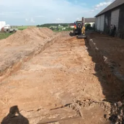 Żółta mini koparka w trakcie wykonywania wykopu liniowego w piaszczystej glebie, widoczny operator, w tle budynek gospodarczy i pole, cień fotografa w dolnej części kadru.