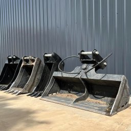 LASKOWSCY Roboty Ziemne Usługi Rolnicze Marcin Laskowski - Rząd używanych, czarnych łyżek do koparko-ładowarki ustawionych przed szarą, metalową halą z otwartymi drzwiami.