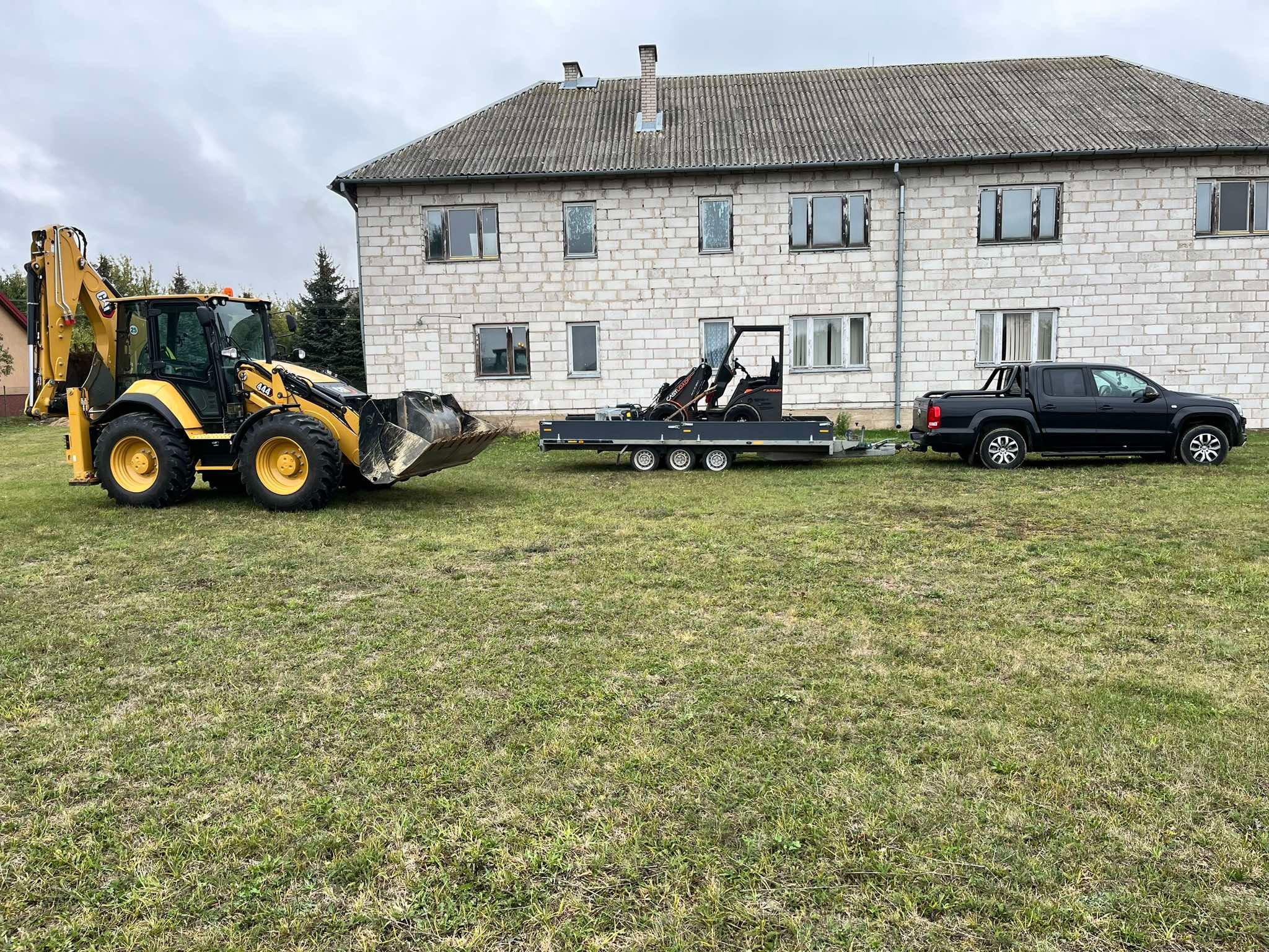 Żółto-czarna koparko-ładowarka CAT 444, czarny pickup Volkswagen Amarok ciągnący przyczepę z miniładowarką na tle budynku z białej cegły i zielonego trawnika.