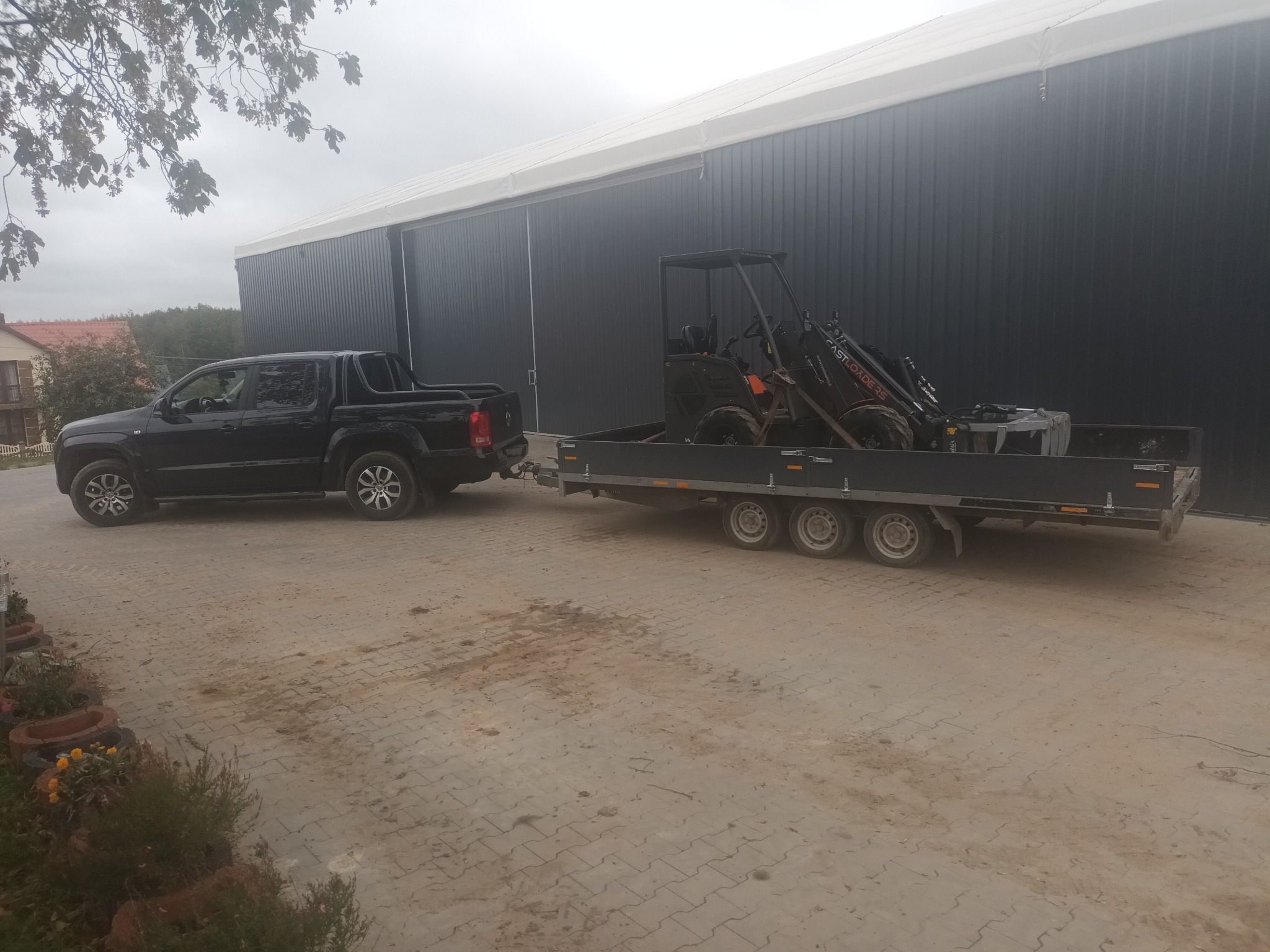 Czarny pick-up Volkswagen Amarok ciągnie przyczepę z mini ładowarką CASTLOADER 35 na tle szarego magazynu z białym dachem.