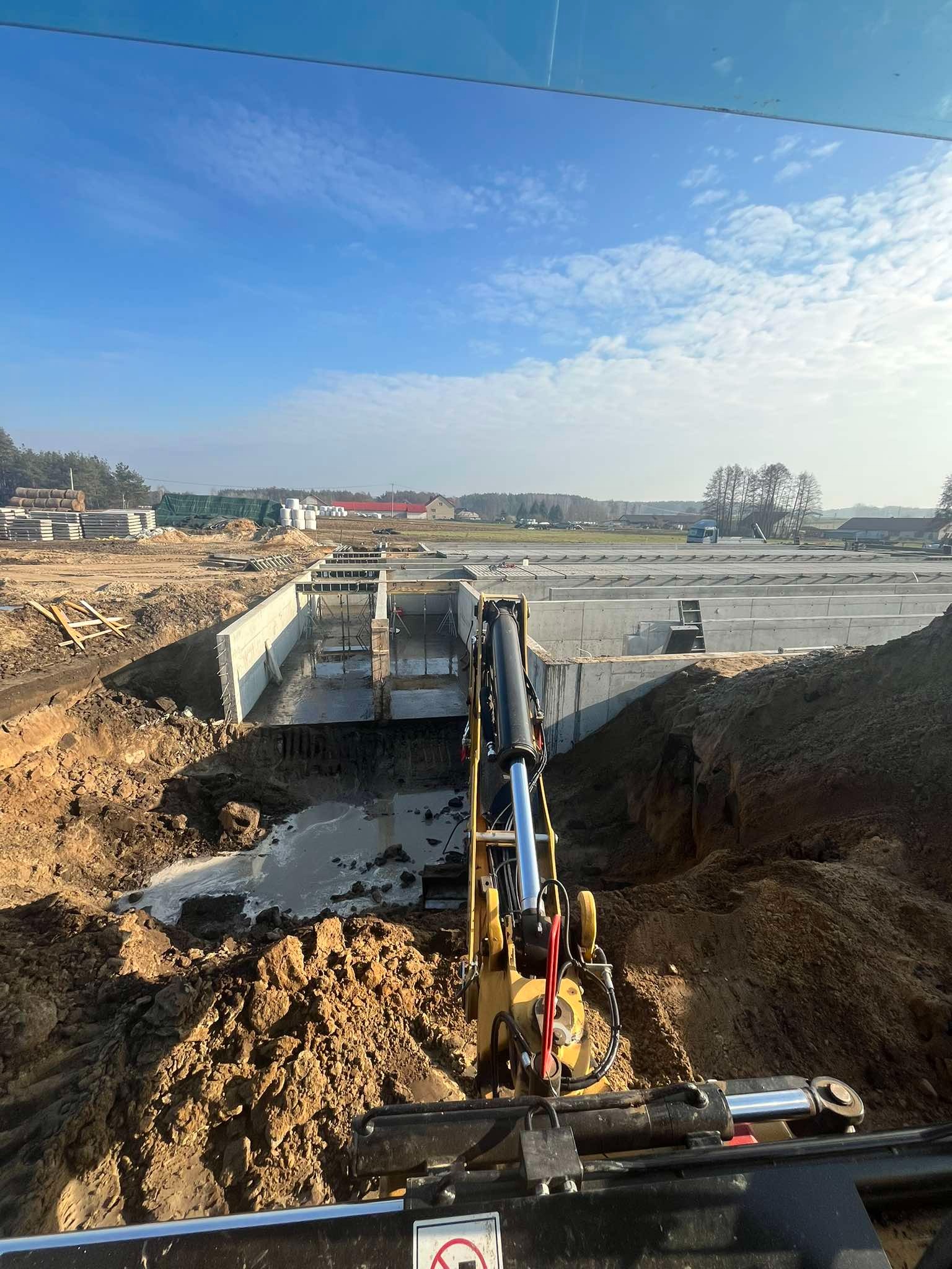 Żółta koparka na pierwszym planie, wykop pod fundamenty z częściowo zalaną wodą, widok z kabiny operatora, błękitne niebo w tle.