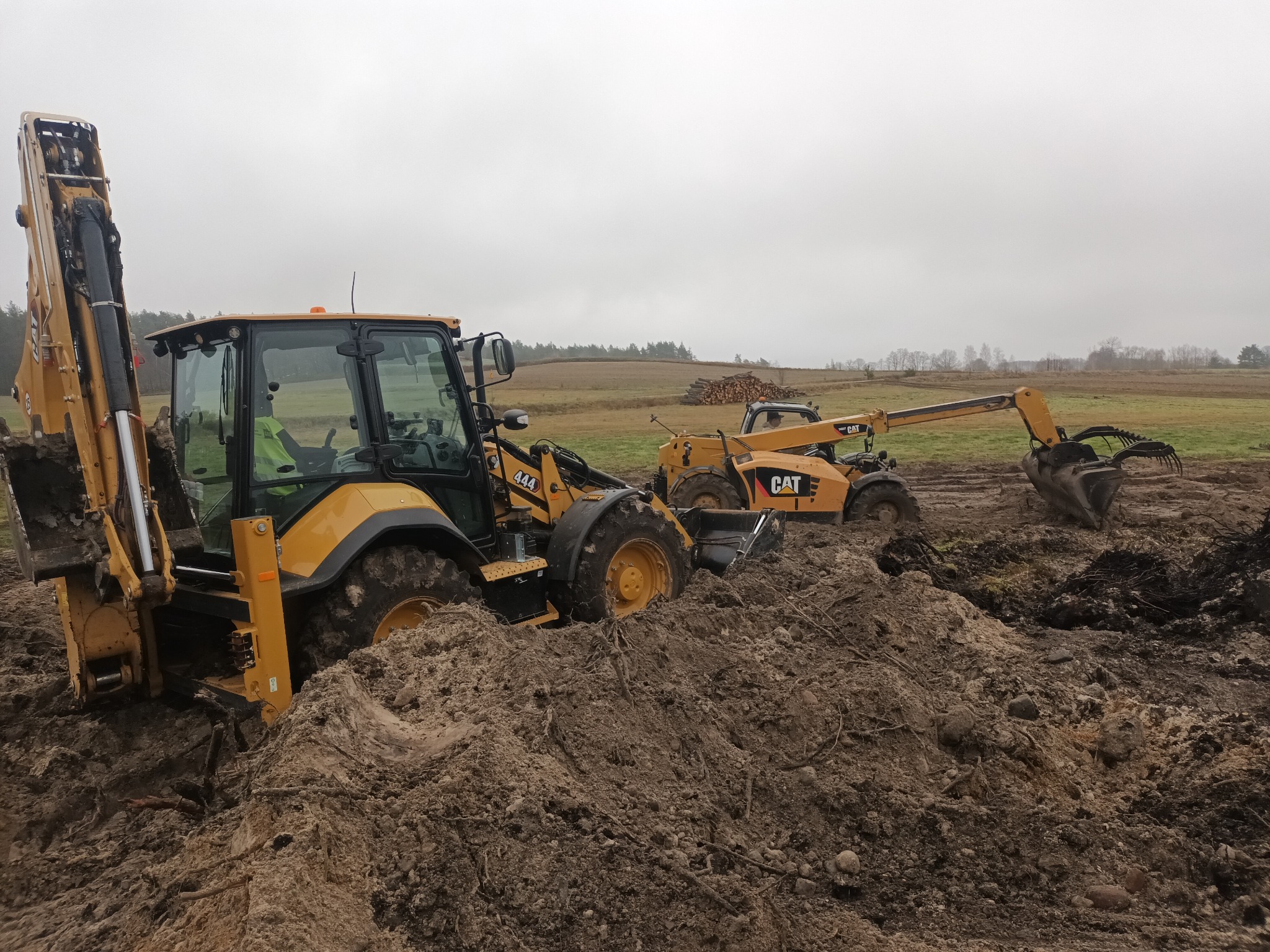 Żółta koparko-ładowarka CAT zagrzebana w błocie podczas prac ziemnych na polu, w tle widoczny drugi pojazd CAT i sterta drewna.