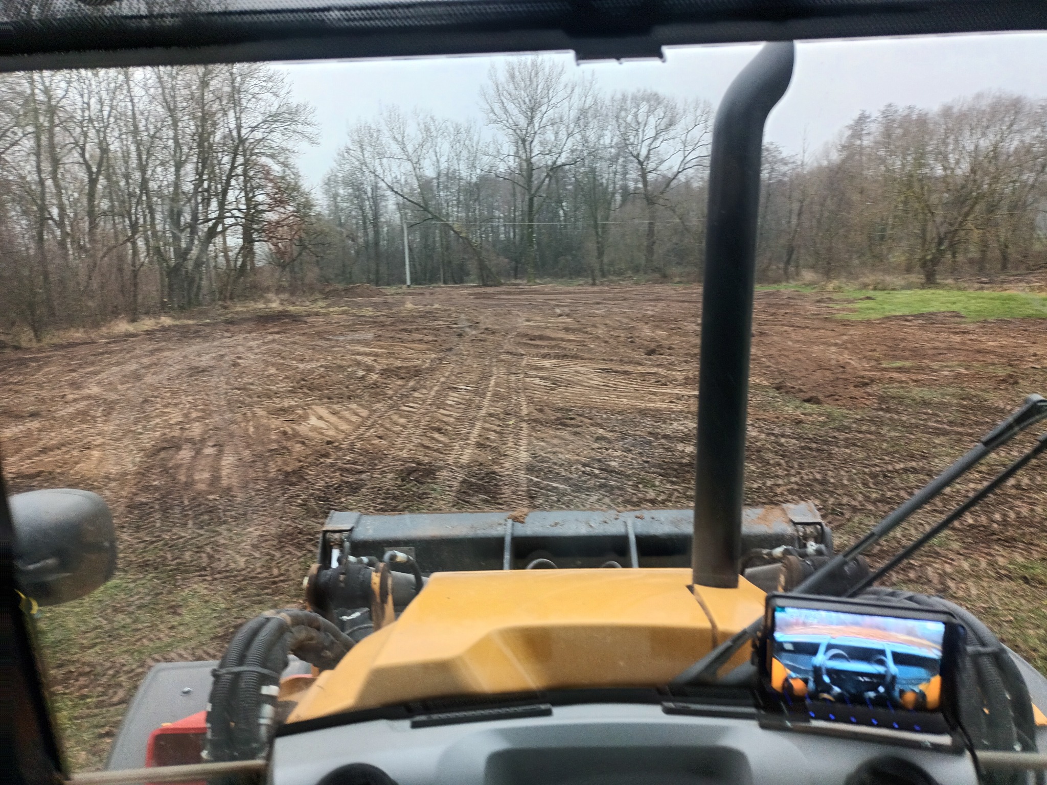 Widok z kabiny koparko-ładowarki na teren po niwelacji, widoczne ślady opon na błotnistym podłożu, w tle drzewa bez liści pod pochmurnym niebem.