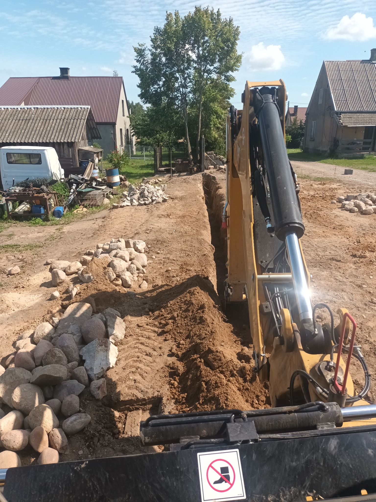 Żółta koparka w trakcie wykonywania wąskiego wykopu w ziemi, w tle zabudowania wiejskie i stos kamieni.